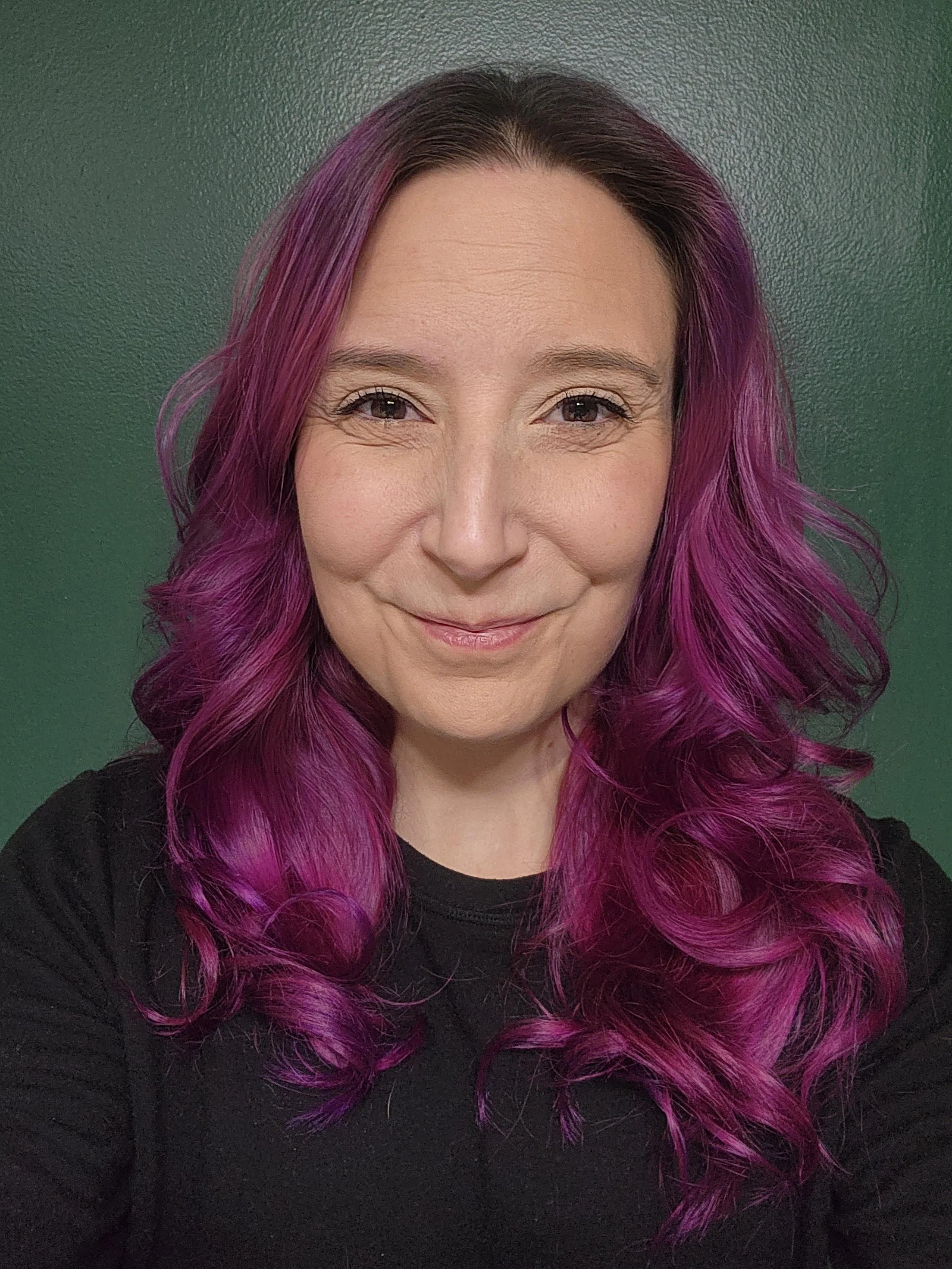 A woman with purple, pink, and magenta hair styled in loose curls, smiling softly with a neutral green background.