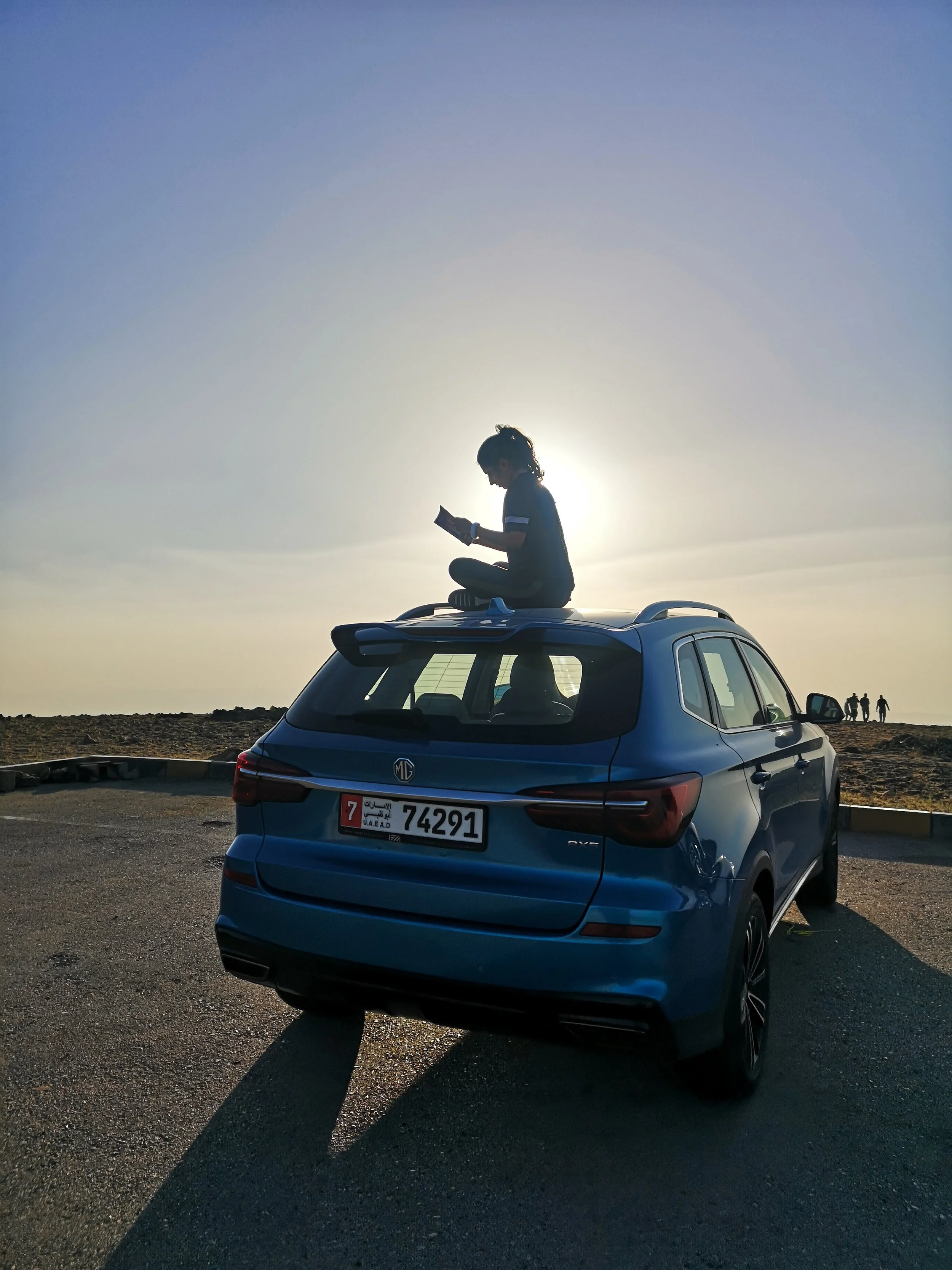 A young girl sitting on the roof of a car, using a tablet, with the sun setting in the background