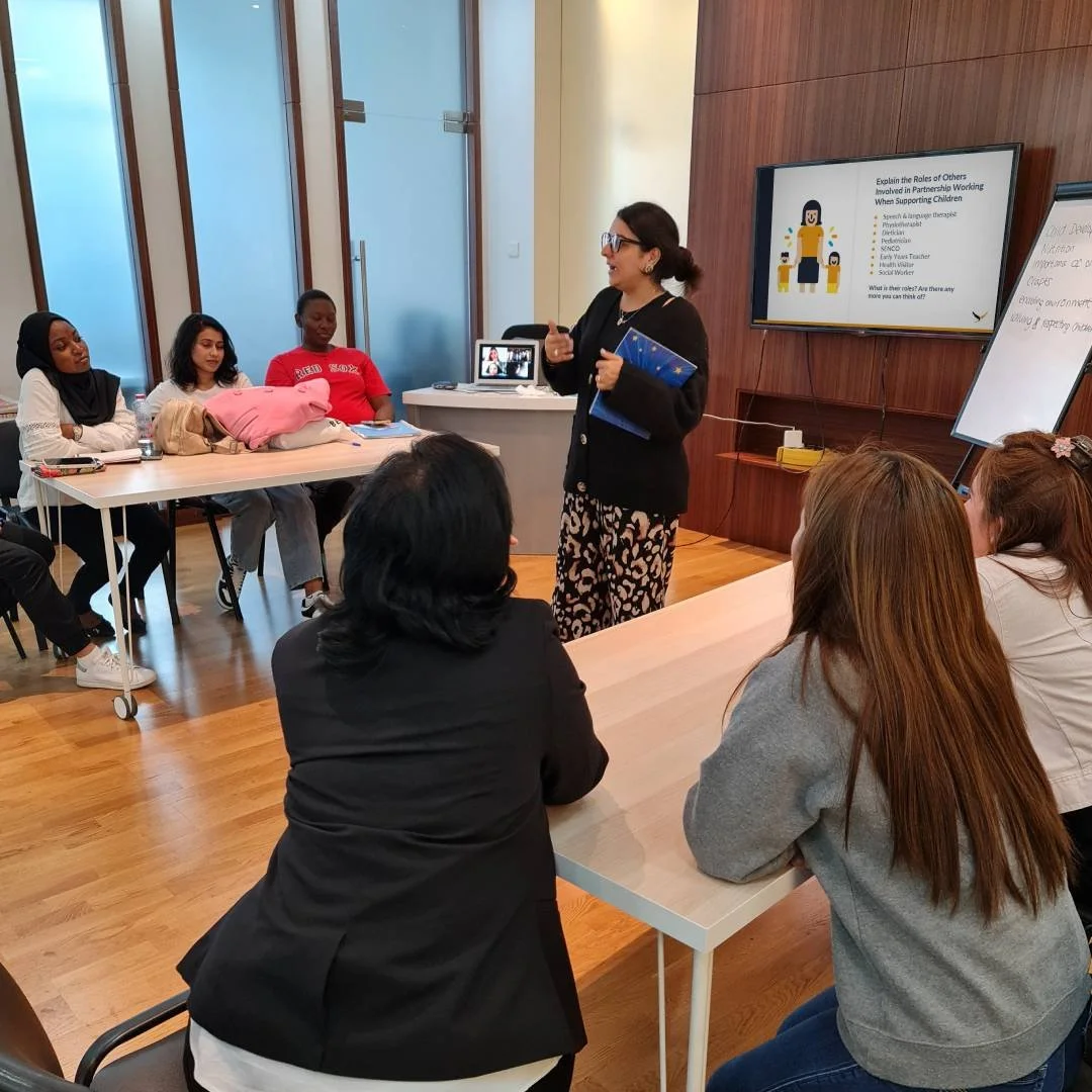 A woman standing and speaking to a group of women seated at a table in a classroom or meeting room, with a large screen behind her displaying a presentation.