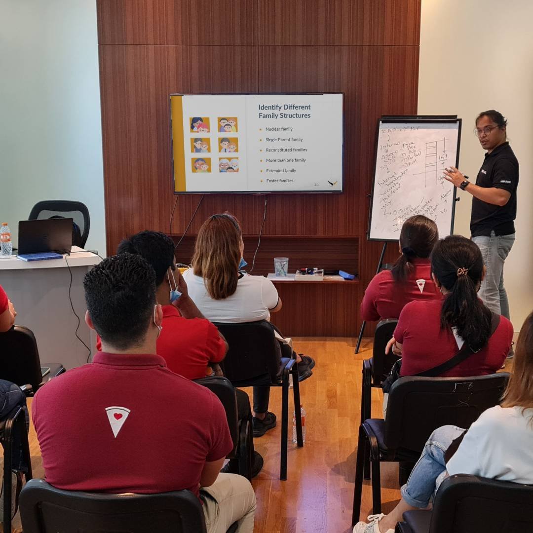 A man giving a lecture to a group of people in a classroom, with a presentation slide on the wall titled 'Identify Different Family Structures' and a whiteboard with notes.