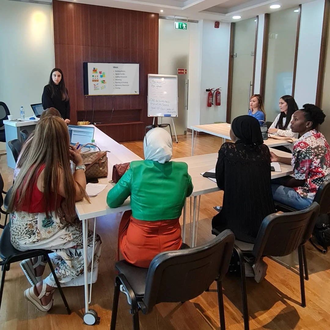 A diverse group of women attending a presentation in a conference room, with a female presenter at the front, screen displaying ideas, and a whiteboard.