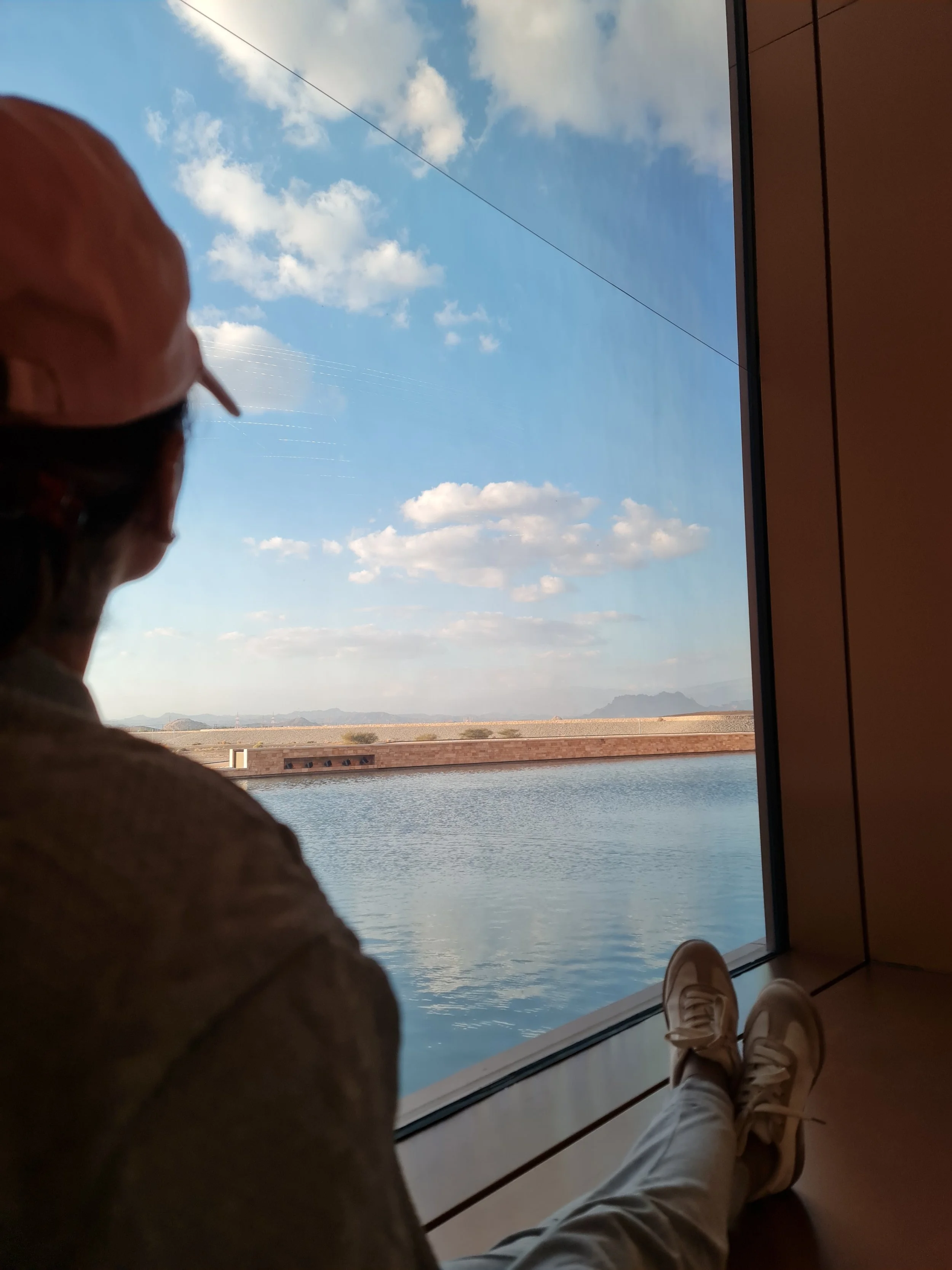 View of a body of water with a shoreline, mountains, blue sky, and clouds seen through a large window. In the foreground, a person wearing a cap, tan pants, and white sneakers is sitting with their legs extended, with their feet visible near the bottom of the image.