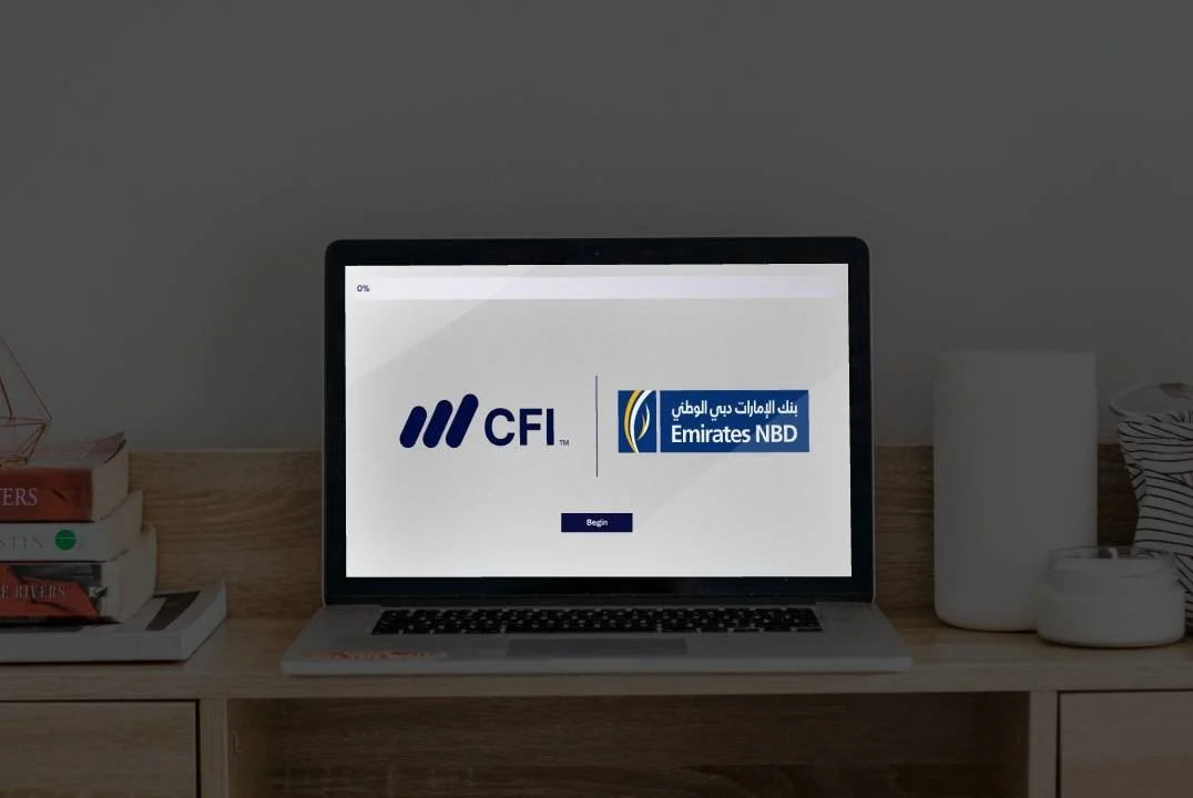 Laptop on a wooden desk displaying the CFI and Emirates NBD logos with a Begin button, surrounded by books and decorative objects.