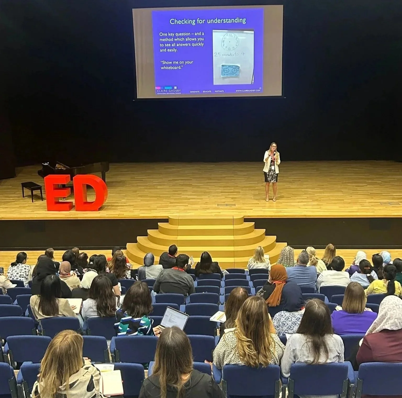 A woman is giving a presentation on a stage in front of a large audience at an education conference. The stage has red letters spelling 'ED' and a grand piano. A screen behind the speaker displays a slide titled 'Checking for understanding,' with text and images about questioning techniques. The audience is seated in rows, attentively listening and some taking notes.