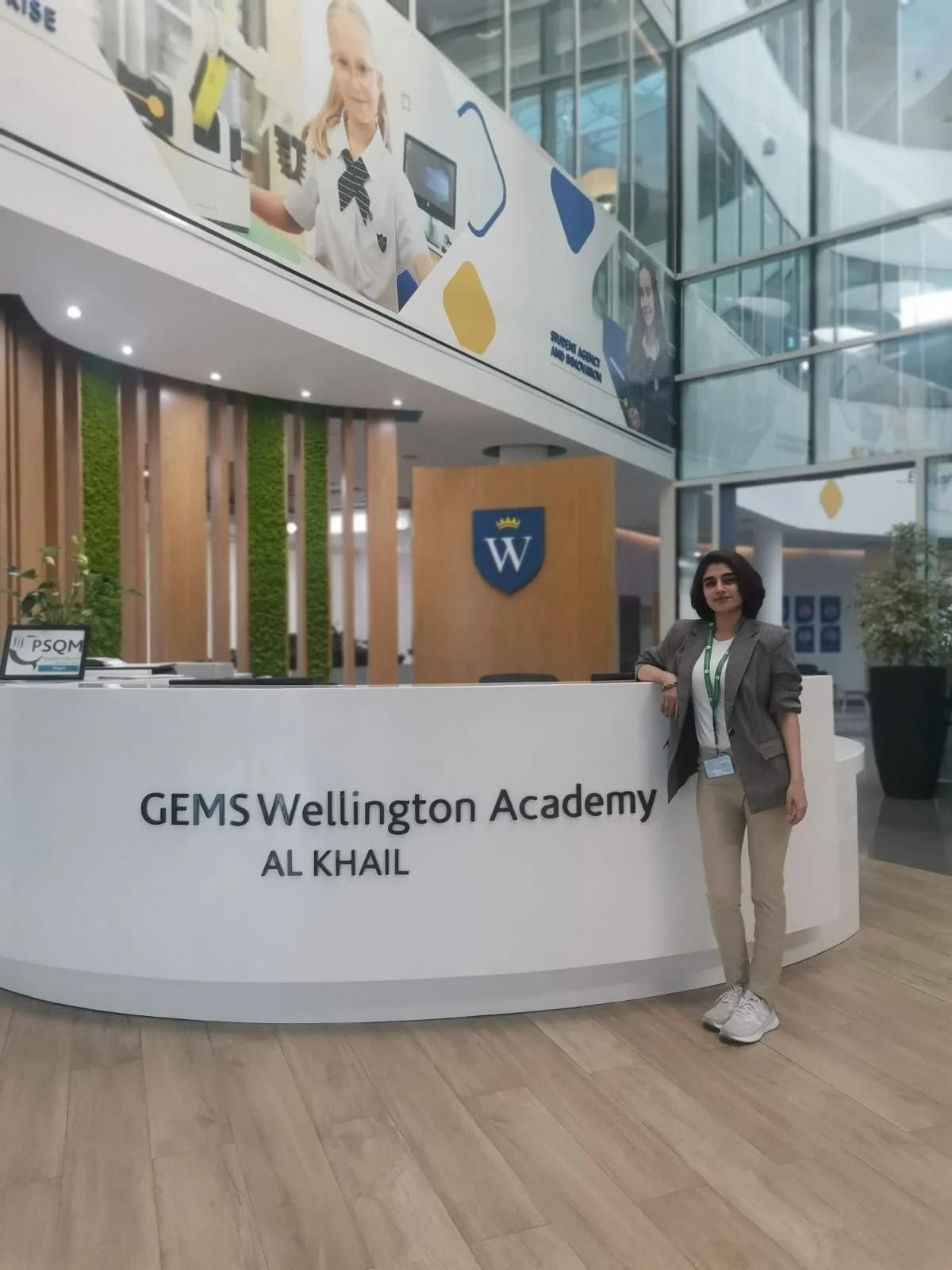 A woman standing at the reception desk of GEMS Wellington Academy in Al Khail, with modern interior and glass architecture in the background.