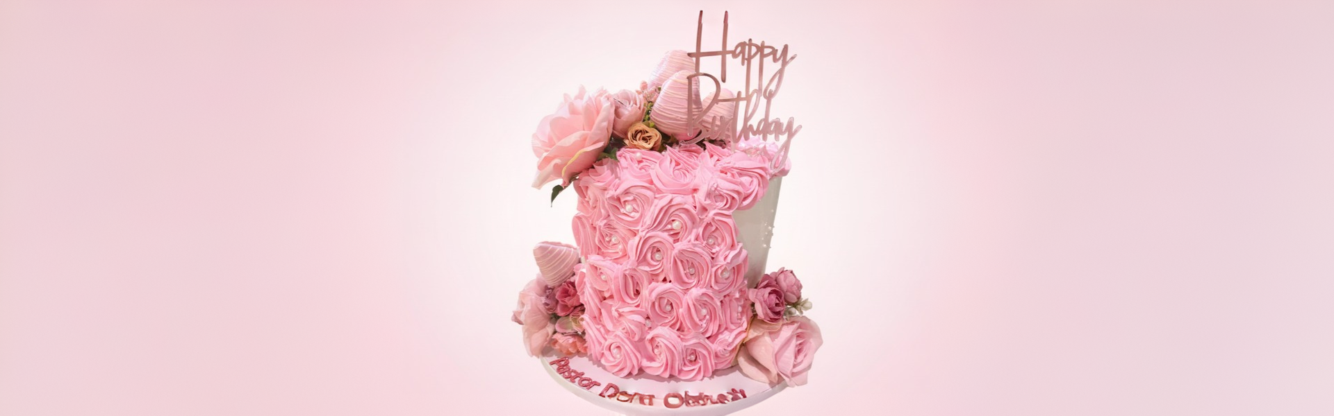 Pink birthday cake decorated with pink roses and rosettes, topped with a "Happy Birthday" cake topper, with pink flowers around the base, against a light pink background.