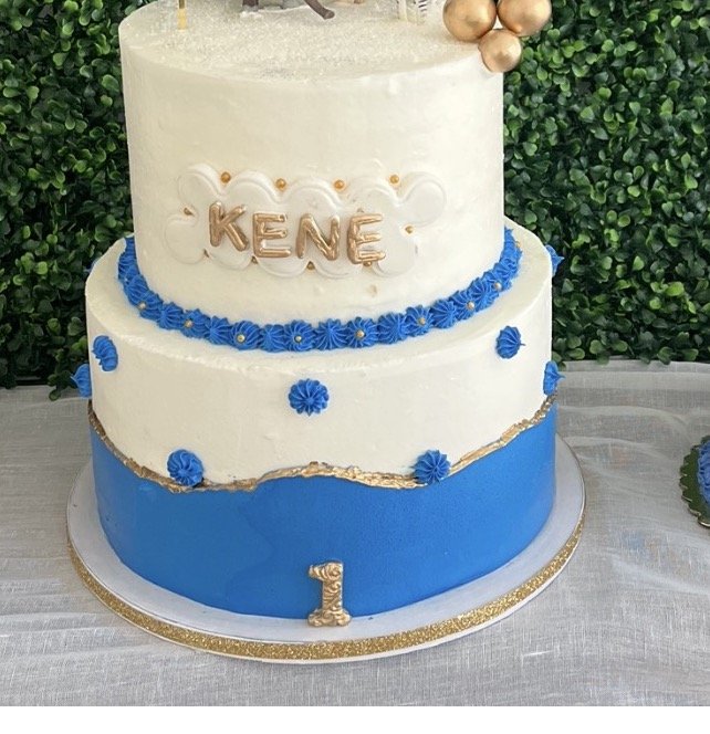 Two-tiered birthday cake with white and blue frosting, decorated with blue flowers and a gold number 1, bearing the name 'KENE' in gold letters on top, set against a green leafy background.