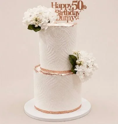A two-tier white birthday cake decorated with white flowers and a topper that says 'Happy 50th Birthday Jenny'.