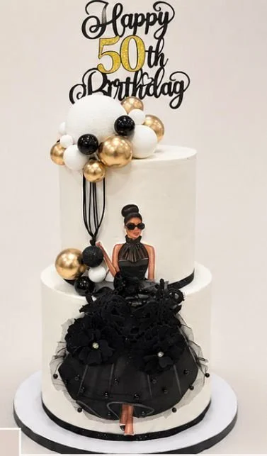 A white birthday cake decorated with black, white, and gold balloons, featuring a woman figurine in a black dress on the front, and a topper that reads 'Happy 50th Birthday' with the number 50 in gold.