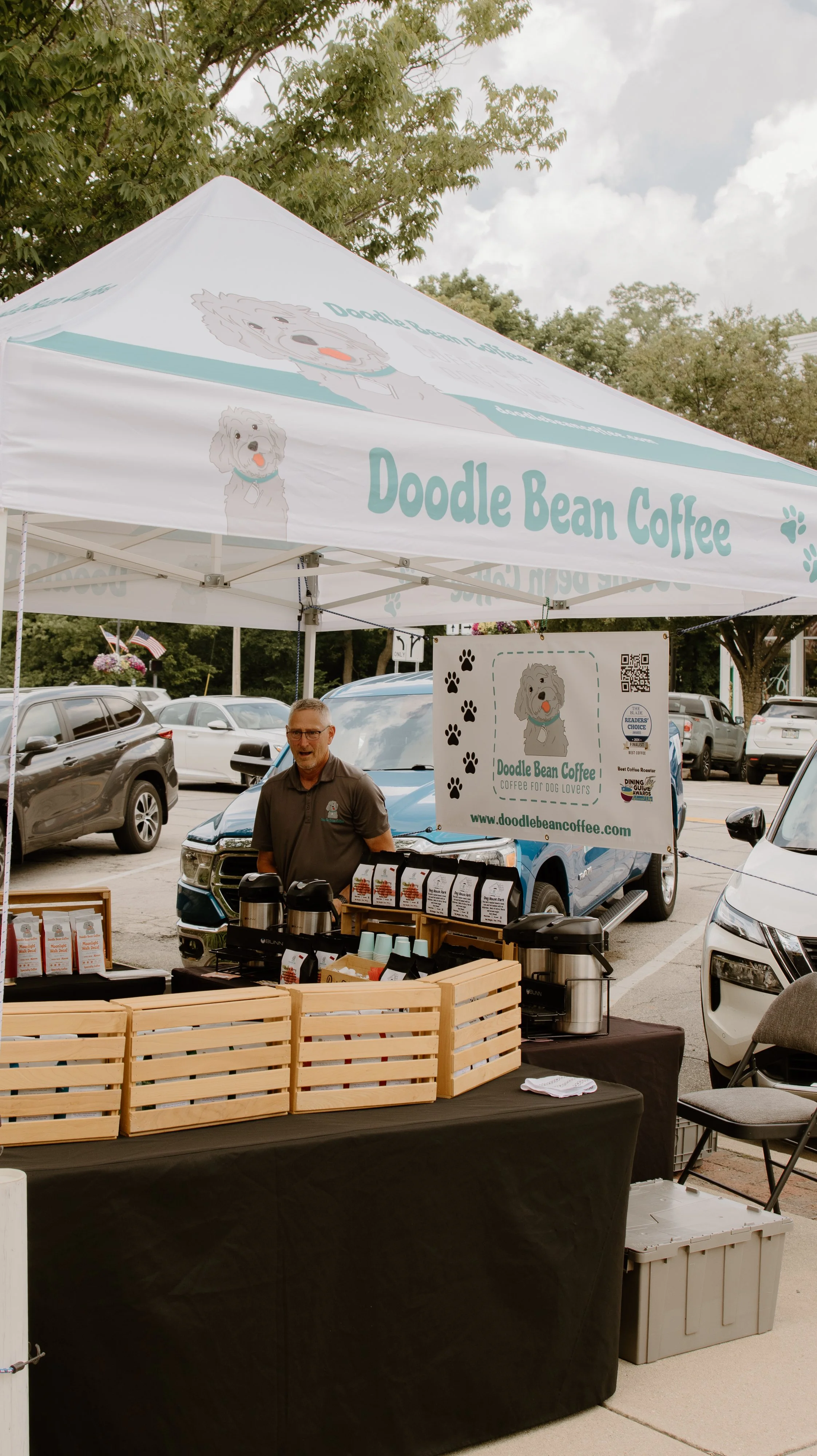 A man standing behind a coffee stand with a sign that reads 'Doodle Bean Coffee' and features a cartoon dog illustration. The stand has various coffee bags, a coffee urn, and cups. In the background, there are parked cars and trees in an outdoor setting.