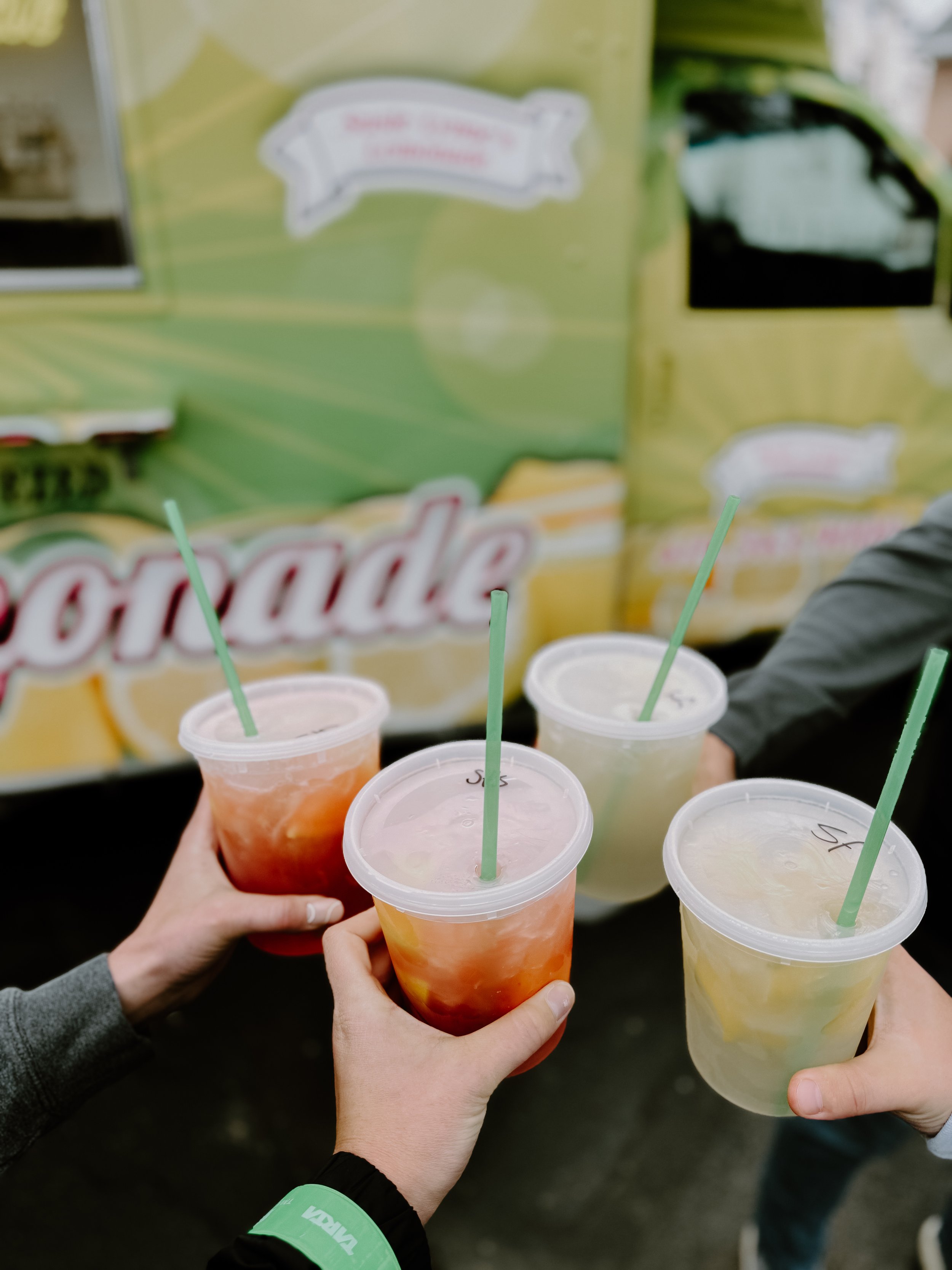 Multiple cups of fruit punches with straws held by different people, with a colorful beverage stand in the background.