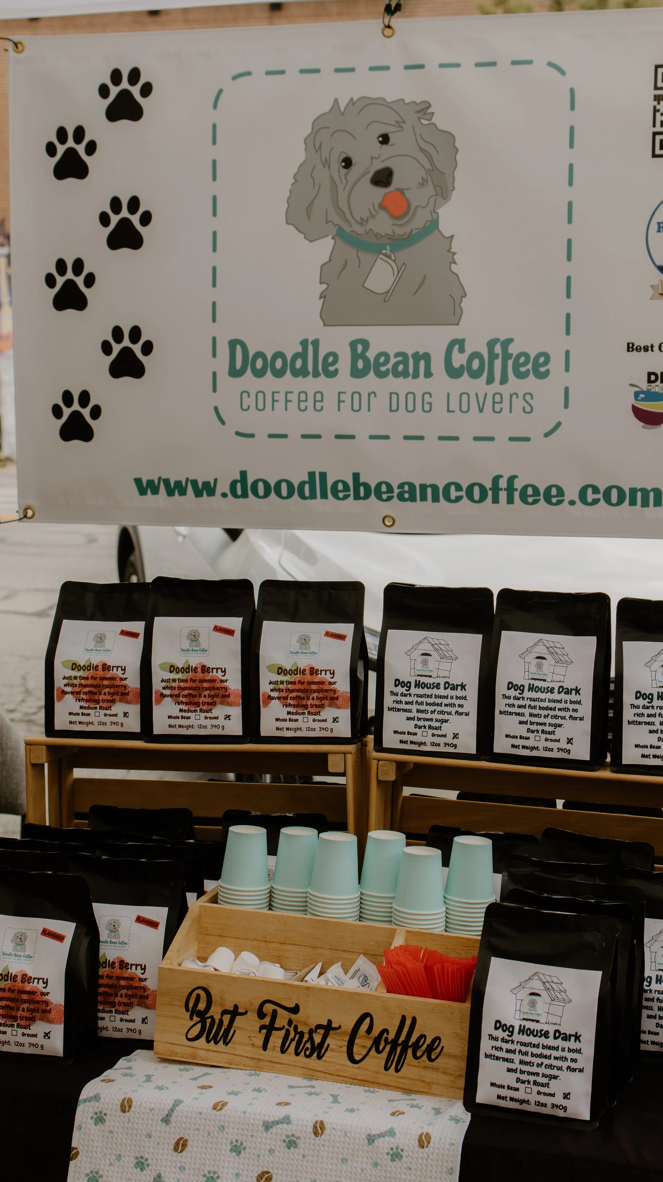 A banner with the logo of Doodle Bean Coffee featuring a cartoon dog with a tongue out and paw prints, advertising coffee for dog lovers. Below, packs of coffee and cups are displayed at a booth, with a sign on a wooden box reading 'But First Coffee'.