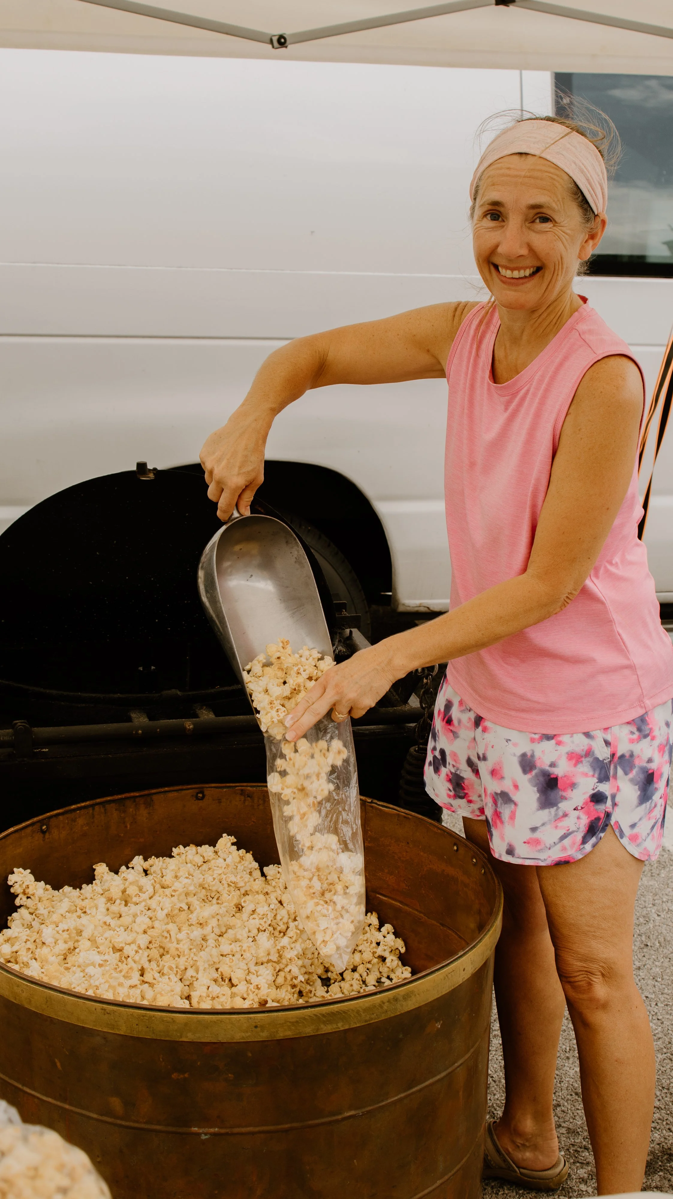 A woman in a pink sleeveless shirt and colorful shorts is pouring popcorn from a large metal container into a plastic bag over a giant metal tub, outdoors.