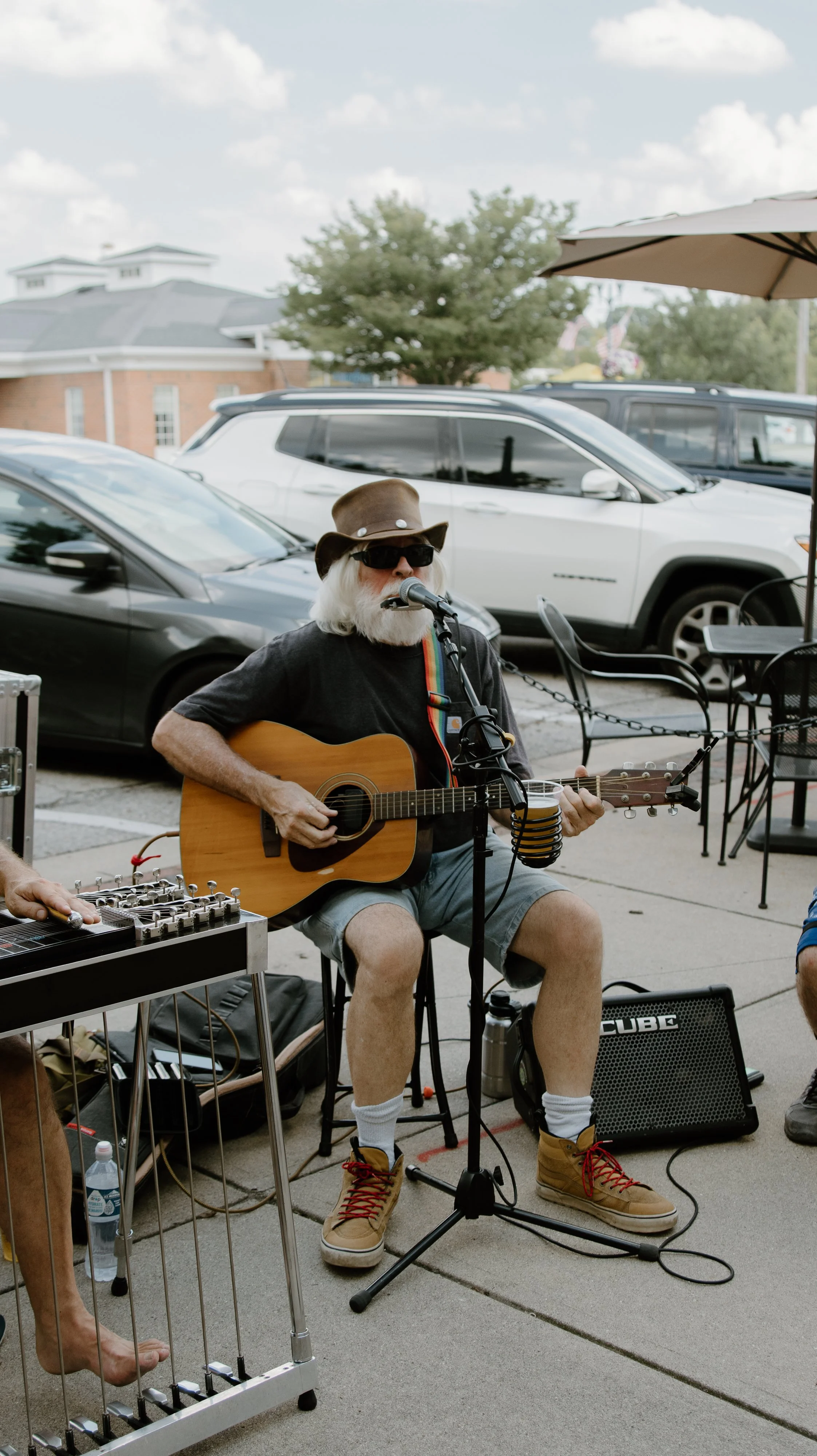 An elderly man with a white beard, wearing sunglasses, a brown hat, a black t-shirt, gray shorts, and tan boots, is playing an acoustic guitar and singing into a microphone. He is seated outdoors on a sidewalk with parked cars and a building in the background.