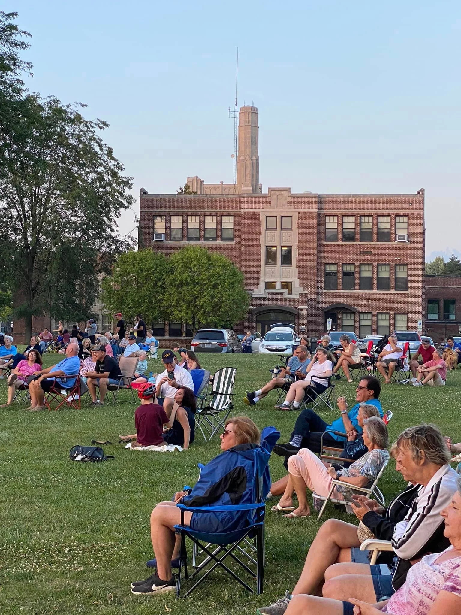 A crowd of people sitting on chairs and on the grass in a park, watching an outdoor event, with a brick building and a tall tower in the background.