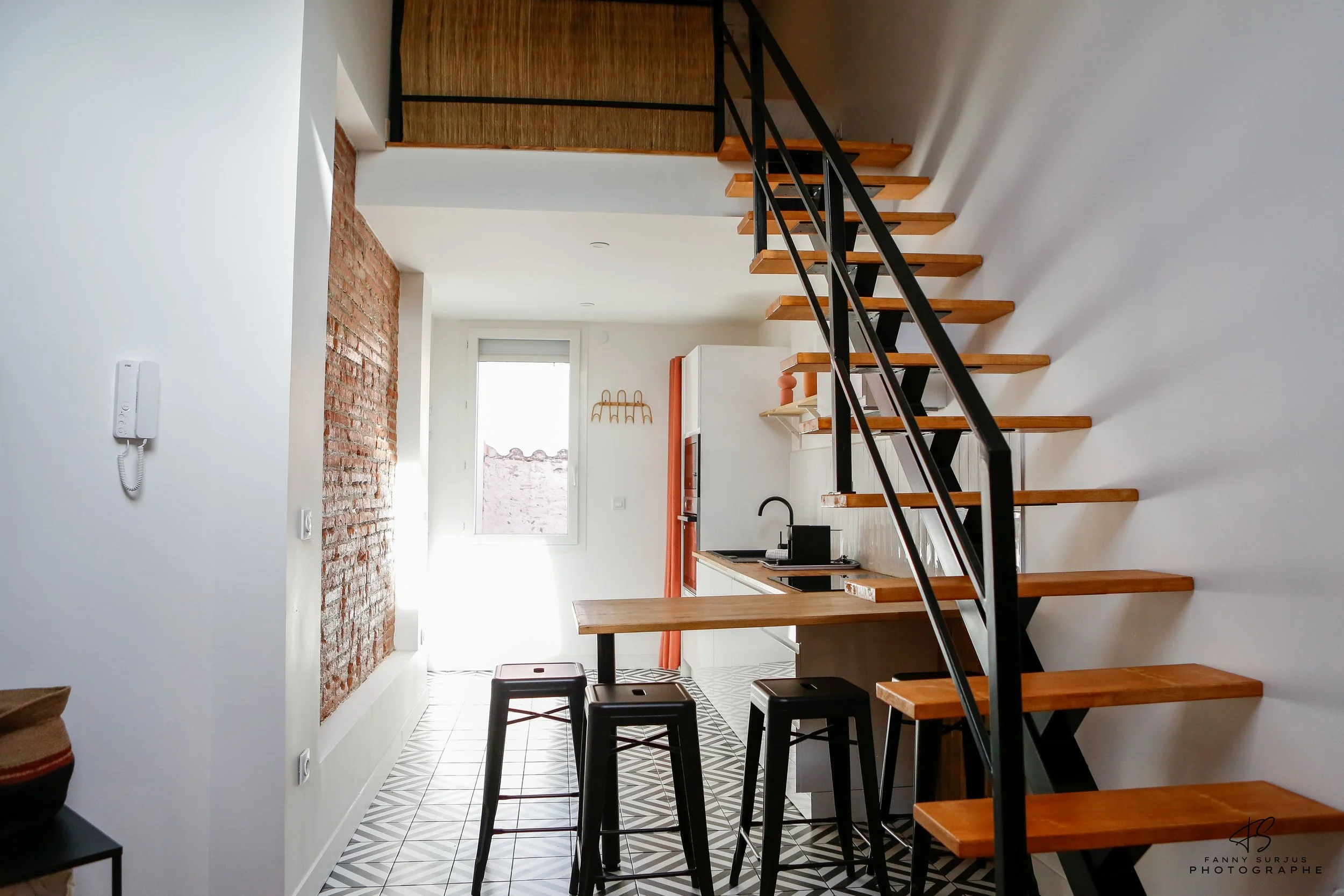 Interior of a modern apartment with a staircase, kitchen area, brick wall, and patterned tile floor.