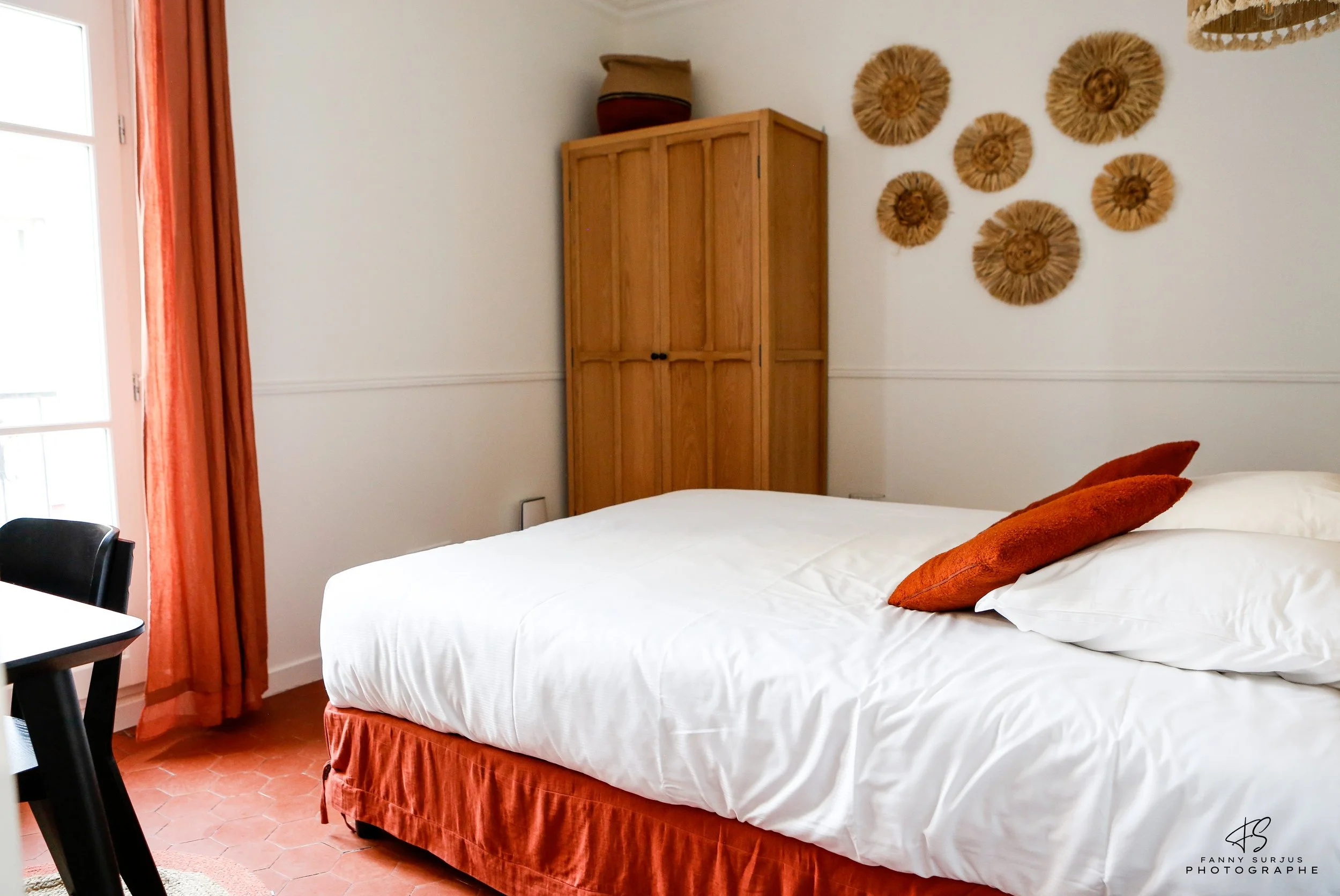A bedroom with a white bed, orange pillows, and an orange bed skirt. There is a wooden wardrobe against the wall, and wall decorations resembling wheat or straw in circular arrangements. An orange curtain covers a window on the left side.