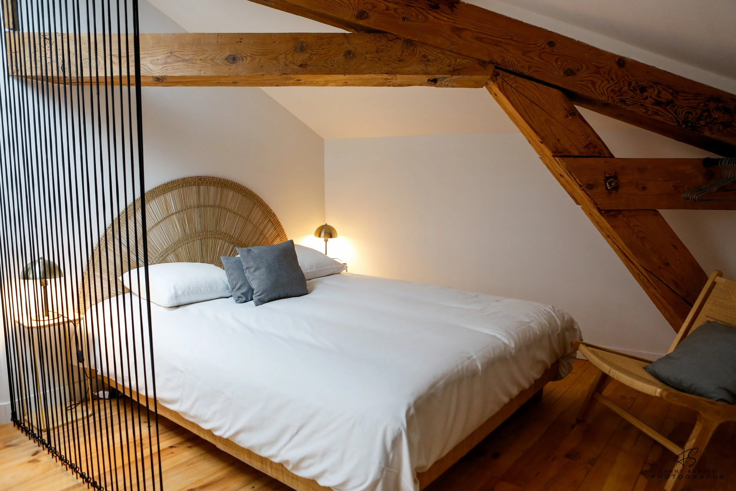 A bedroom with a sloped ceiling, featuring a bed with white linens and white and black pillows. There is a woven headboard, a small bedside lamp, and a wooden chair with a cushion. The room has exposed wooden beams and a wooden floor.