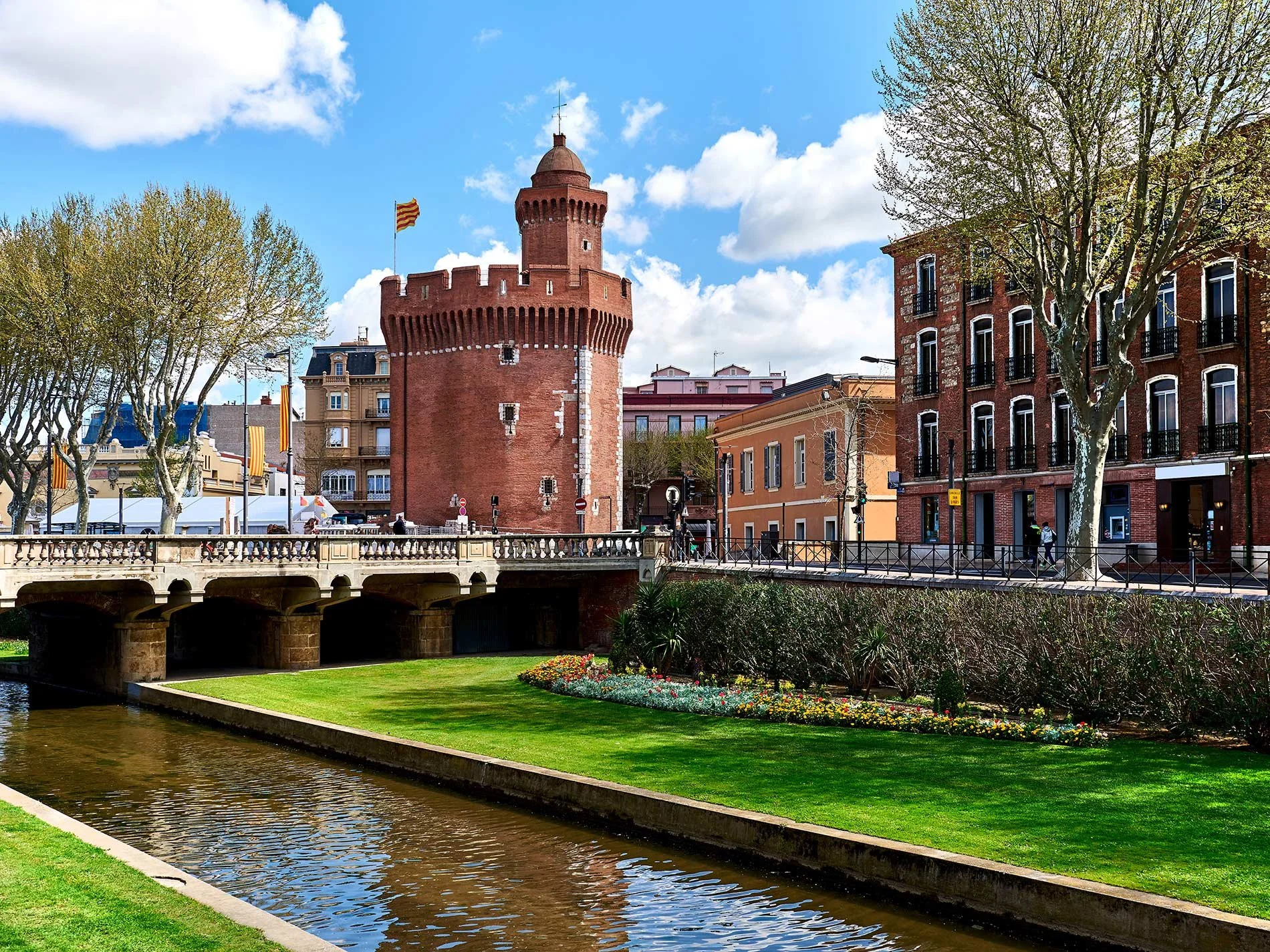A historic red brick tower with a flag called the Castillet in Perpignan, surrounded by buildings, trees, a bridge, and a canal in the with a blue sky.