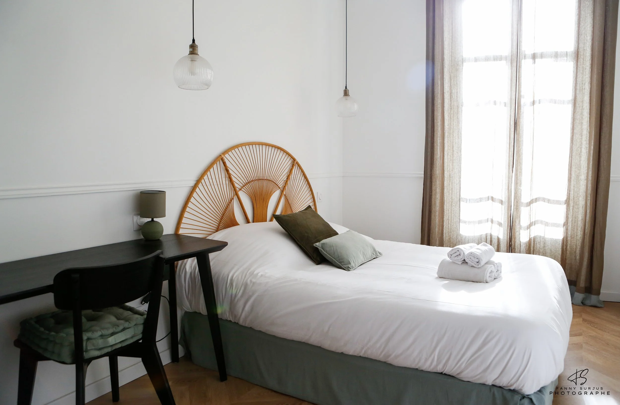 A tidy bedroom with a white wall, two hanging pendant lights, a wooden headboard, two rolled towels on a bed with brown and gray pillows, a small green table lamp, a black desk, and a window with green curtains letting in natural light.