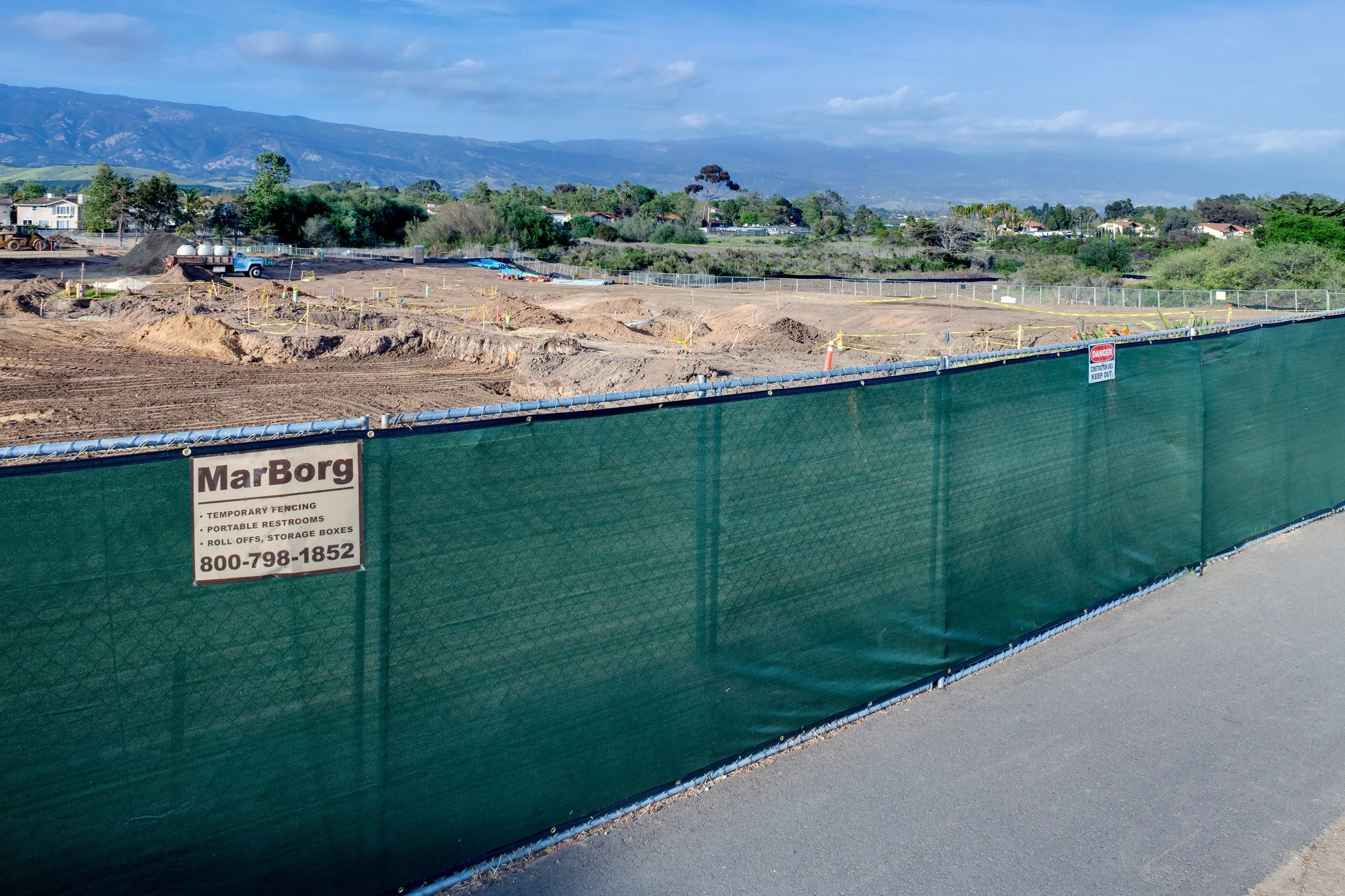 View of a construction site with dirt piles, machinery, and safety fencing, with a mountainous landscape and houses in the background.