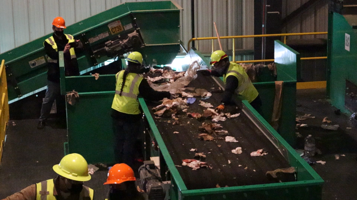Workers wearing high-visibility vests, helmets, and masks sorting meat on a conveyor belt in a processing facility.