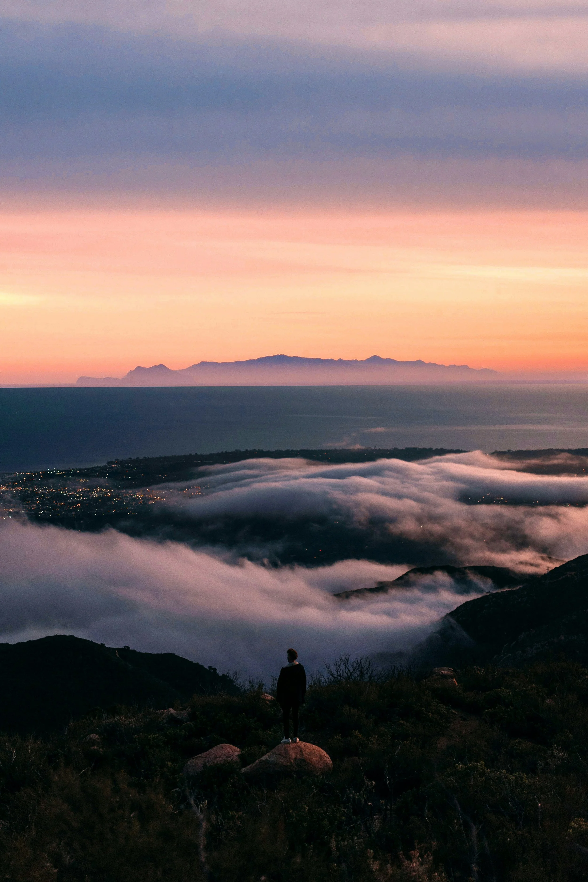 A person standing on a rocky hill at sunset, overlooking a landscape with fog-covered valleys, distant mountains, and a pink and purple sky.