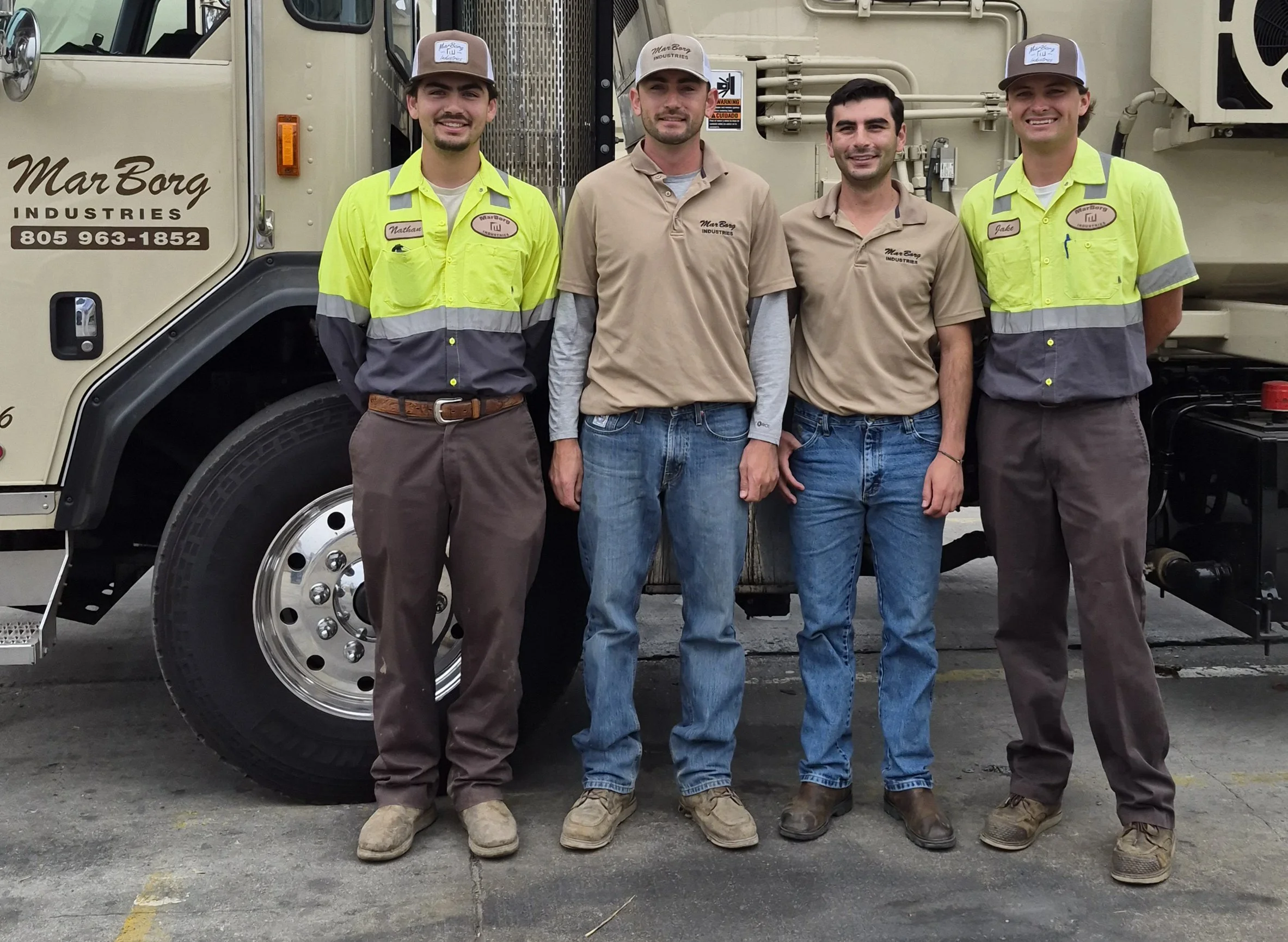 Four workers standing in front of a large industrial vehicle with the company name MarBorg Industries and phone number displayed on it. Two workers are wearing yellow high-visibility shirts, two are wearing beige shirts, and three are wearing hats. They are smiling and posing for the photo.