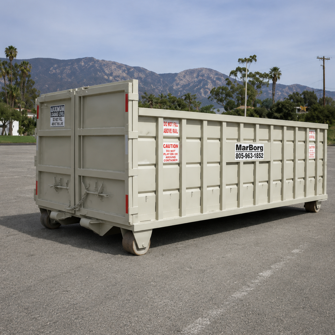 A large beige roll-off trash dumpster with signage for MarBorg Industries, situated in an empty parking lot with mountains and trees in the background under a partly cloudy sky.