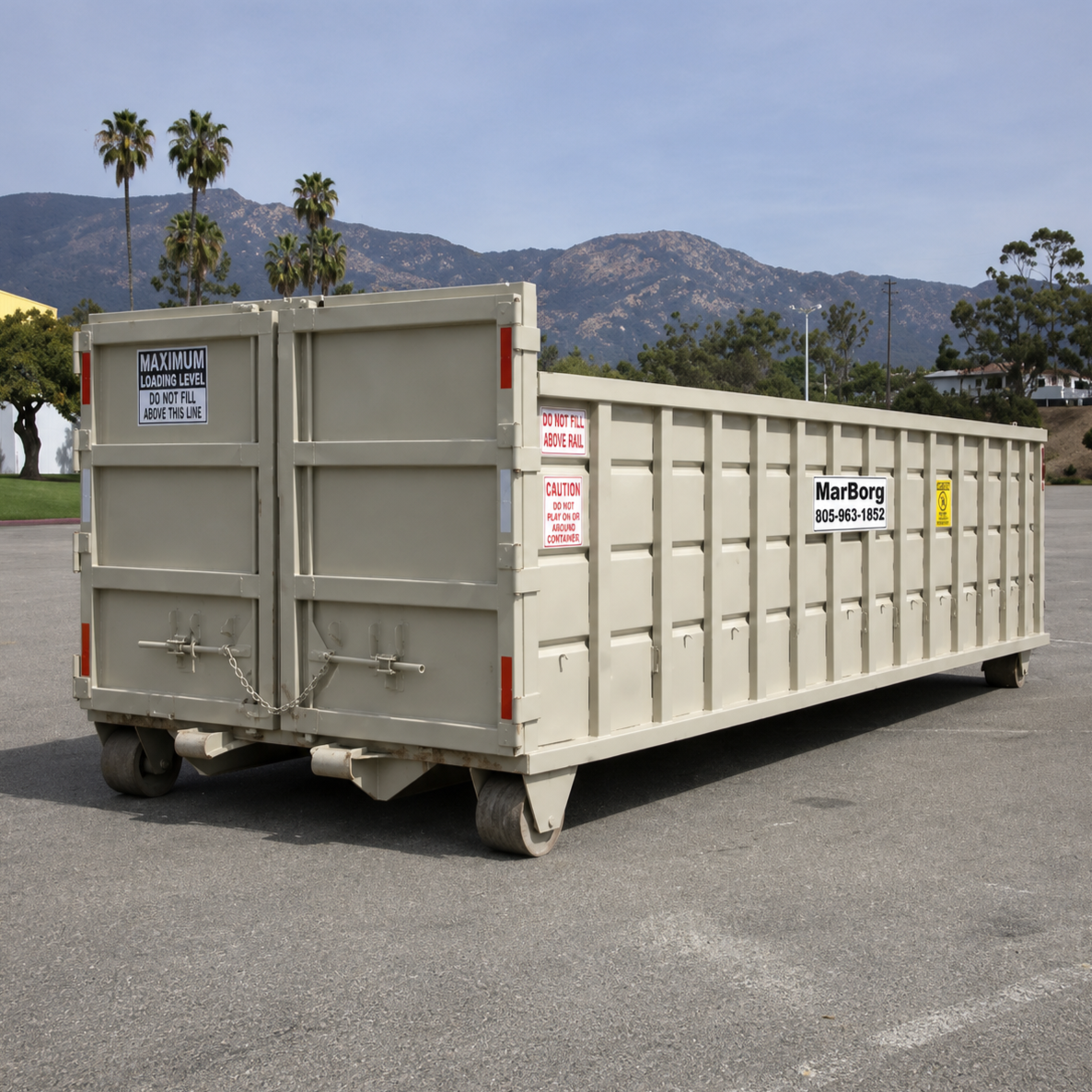 Empty beige roll-off container on a parking lot with mountains, trees, and palm trees in the background and cloudy sky.