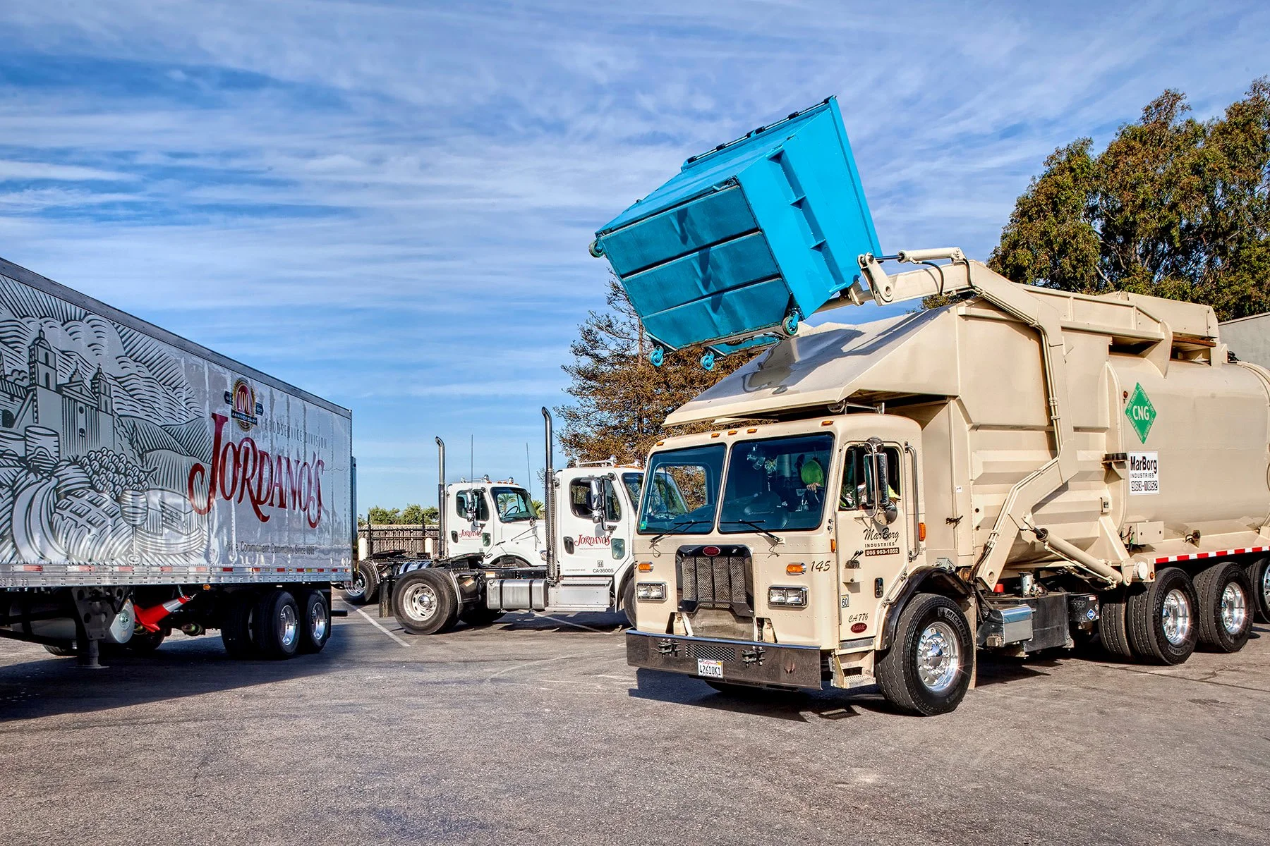 A large MarBorg commercial front-loader truck is emptying a blue 4 yard bin.  There is a Jordano's truck in a parking lot, with other trucks and trees in the background under a partly cloudy sky.