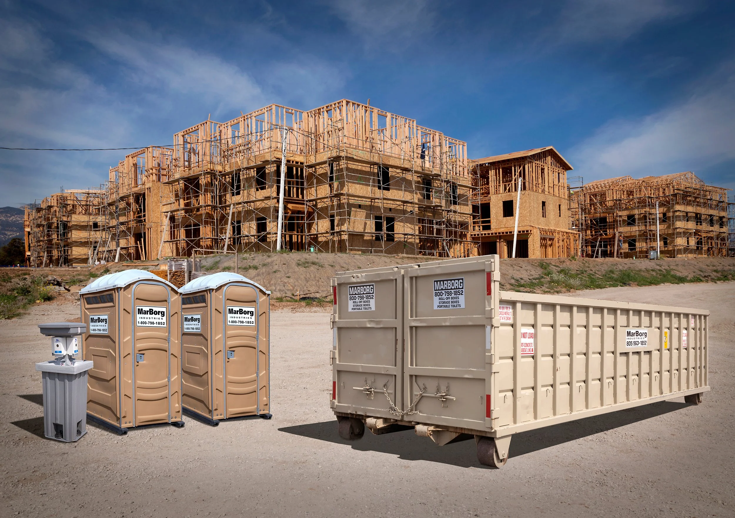Construction site with unfinished wooden buildings, scaffolding, portable toilets, and construction equipment under a partly cloudy sky.