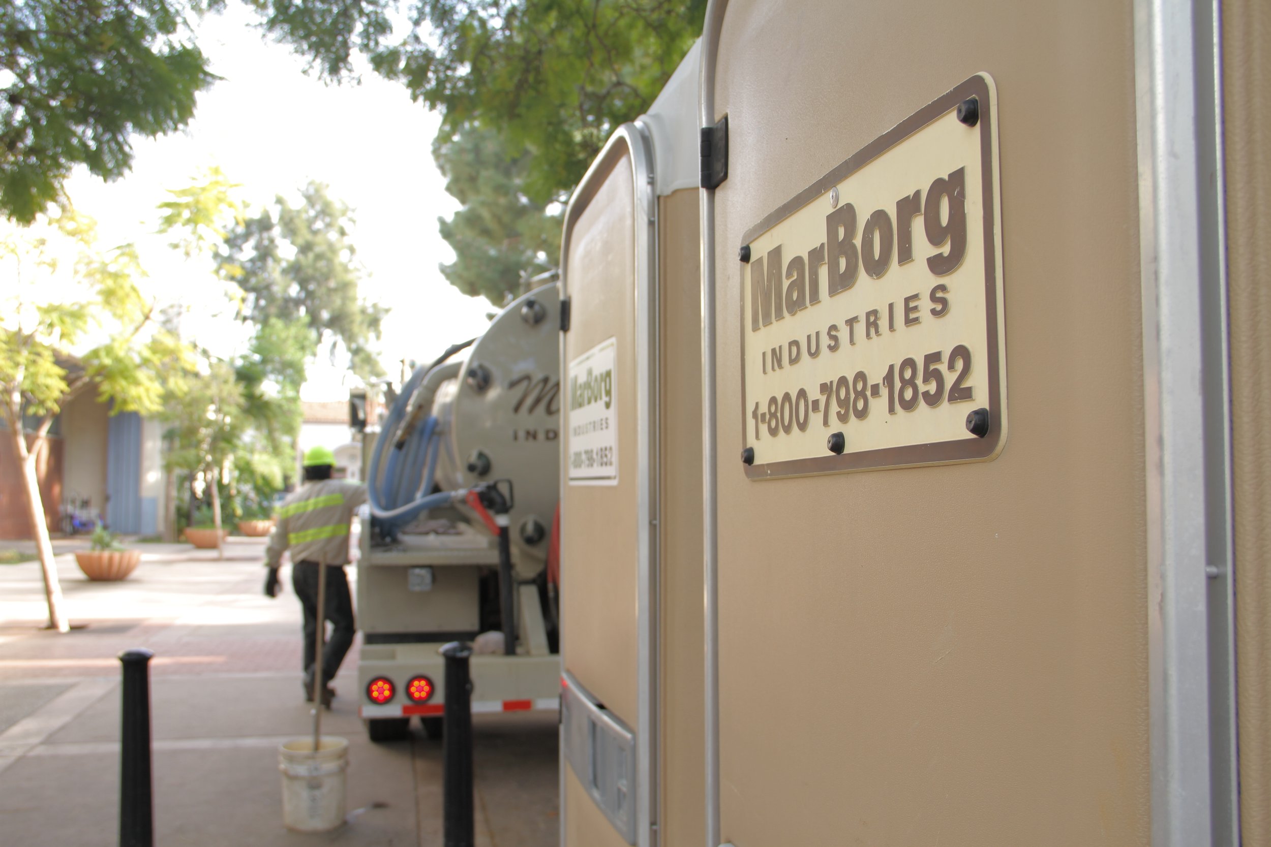 Close-up of a beige portable restroom with a metal sign that reads 'MarBorg Industries' and a phone number, with a street scene and trees in the background.