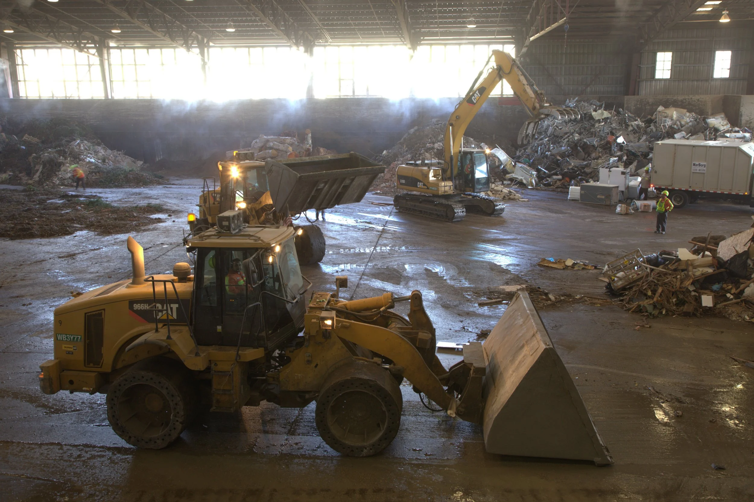 Inside a large construction and demolition recycling facility, construction workers operate heavy machinery, including a front loader and an excavator, as they manage a large pile of mixed debris, scrapyard materials, and waste.