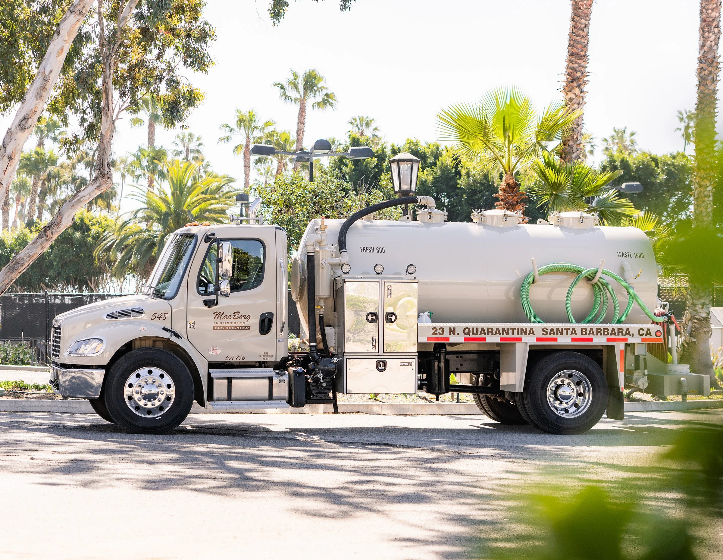 A septic service truck with a white cab and tank, palm trees in the background, and a small green plant partially covering the lower right corner of the image.