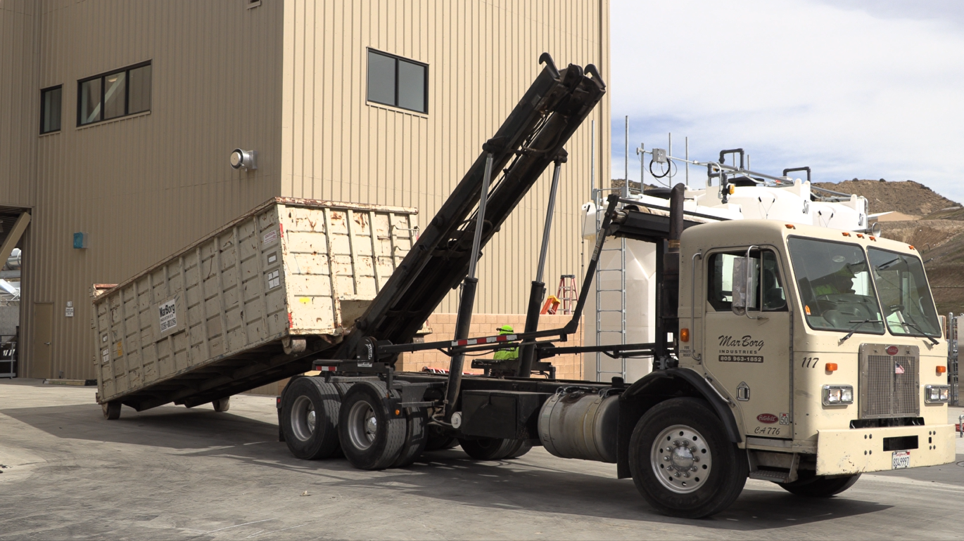 A tow truck with a dumpster attached to its lift arm parked outside a beige industrial building.