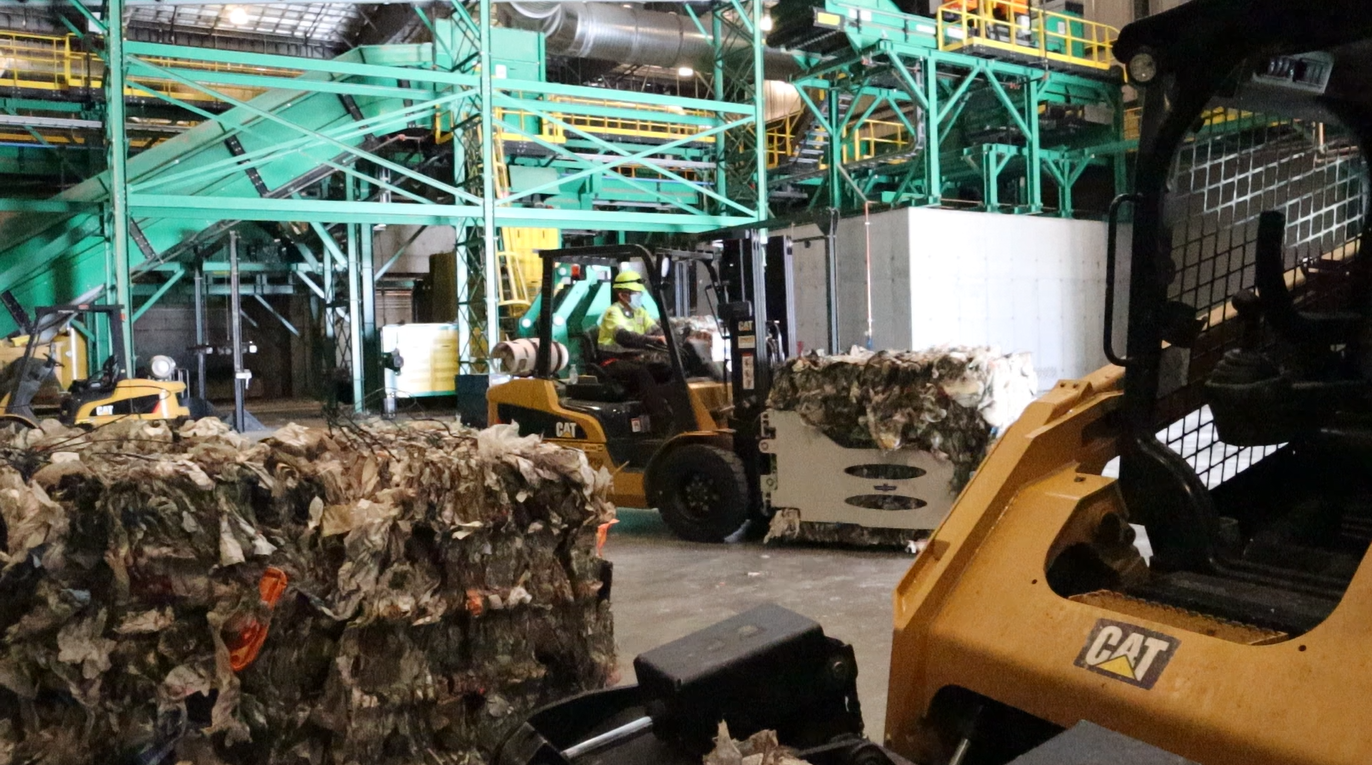 Industrial recycling facility with large machinery and compactors processing paper or plastic waste, with workers operating equipment.