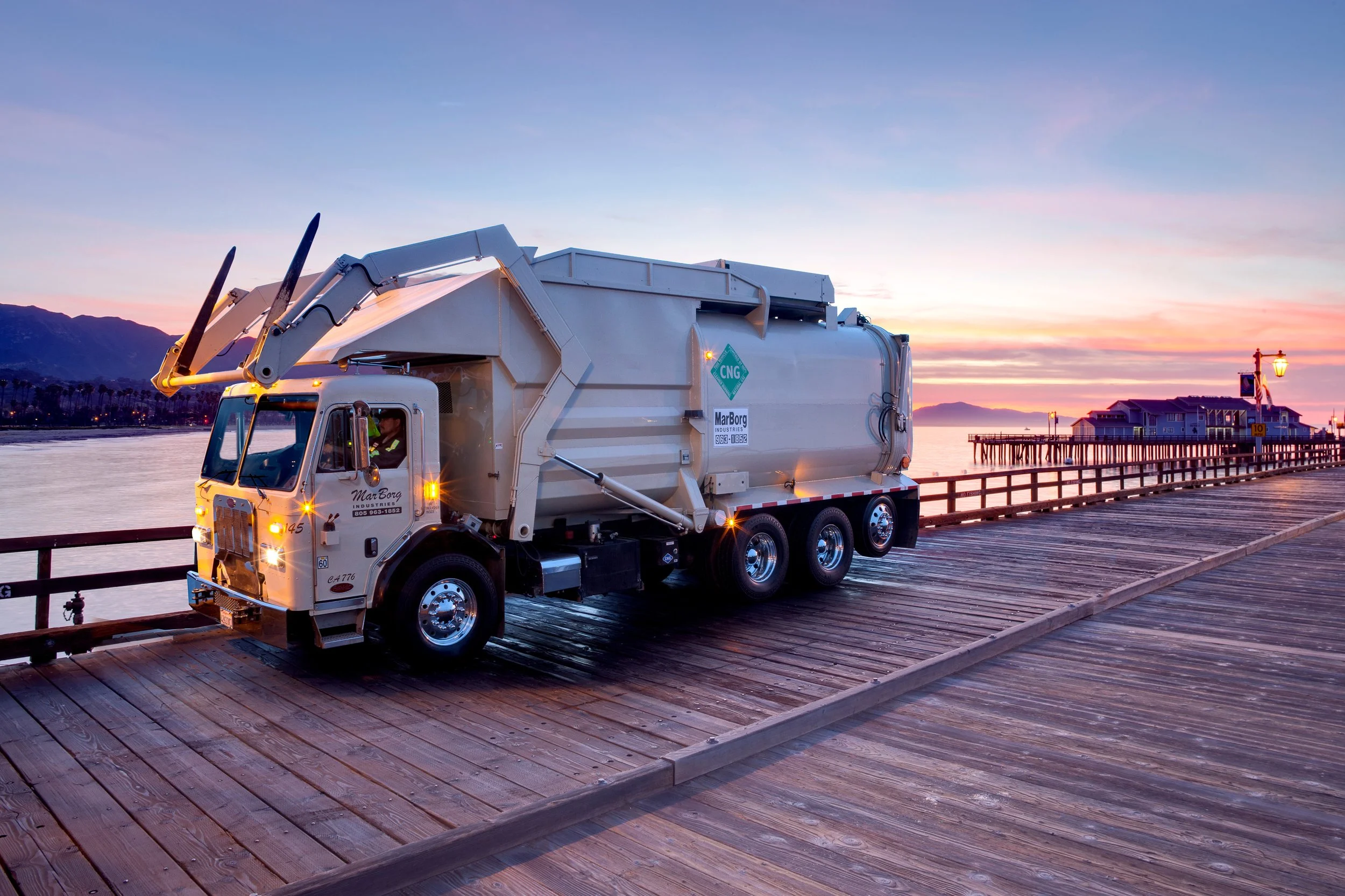 A large white CNG-powered front-loading trash truck parked on the Santa Barbara pier at sunrise, with mountains and a small building in the background.