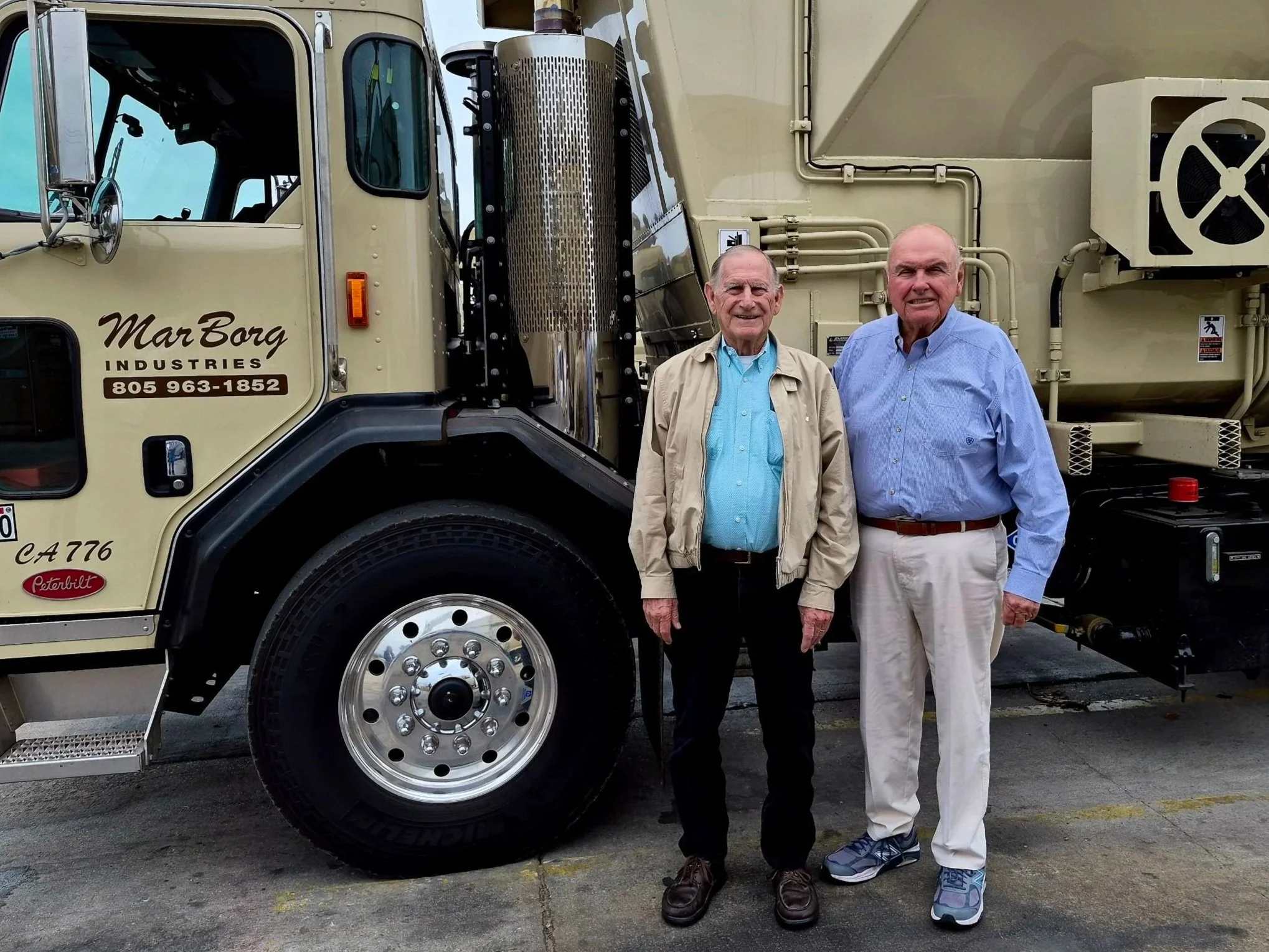 Two elderly men standing in front of a large beige industrial vehicle, possibly a fire truck, with one man wearing a light beige jacket and the other in a light blue shirt.