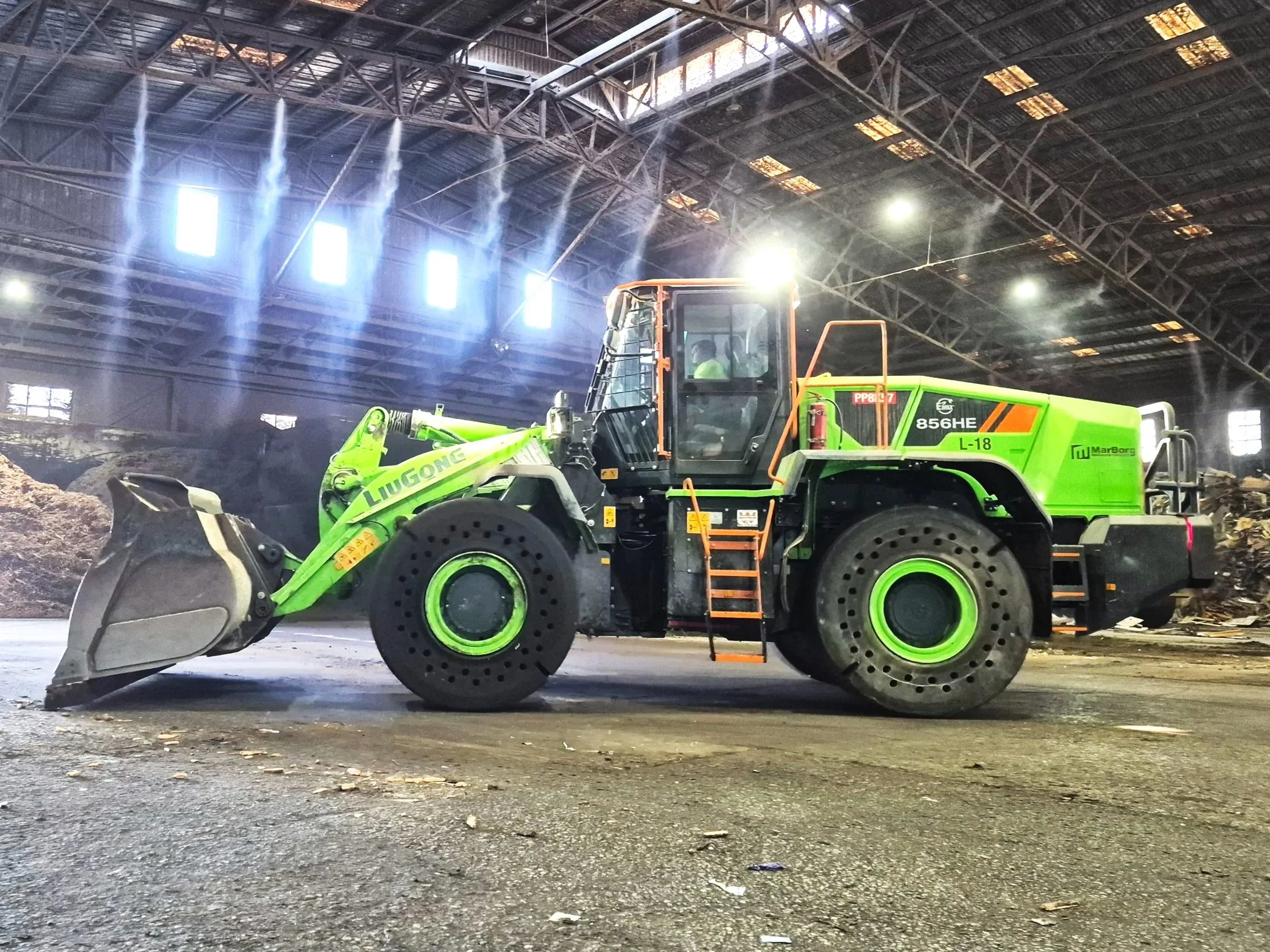A large green and orange front loader construction vehicle inside a spacious warehouse or industrial building, with sunlight streaming through high windows and dust or particles visible in the air.