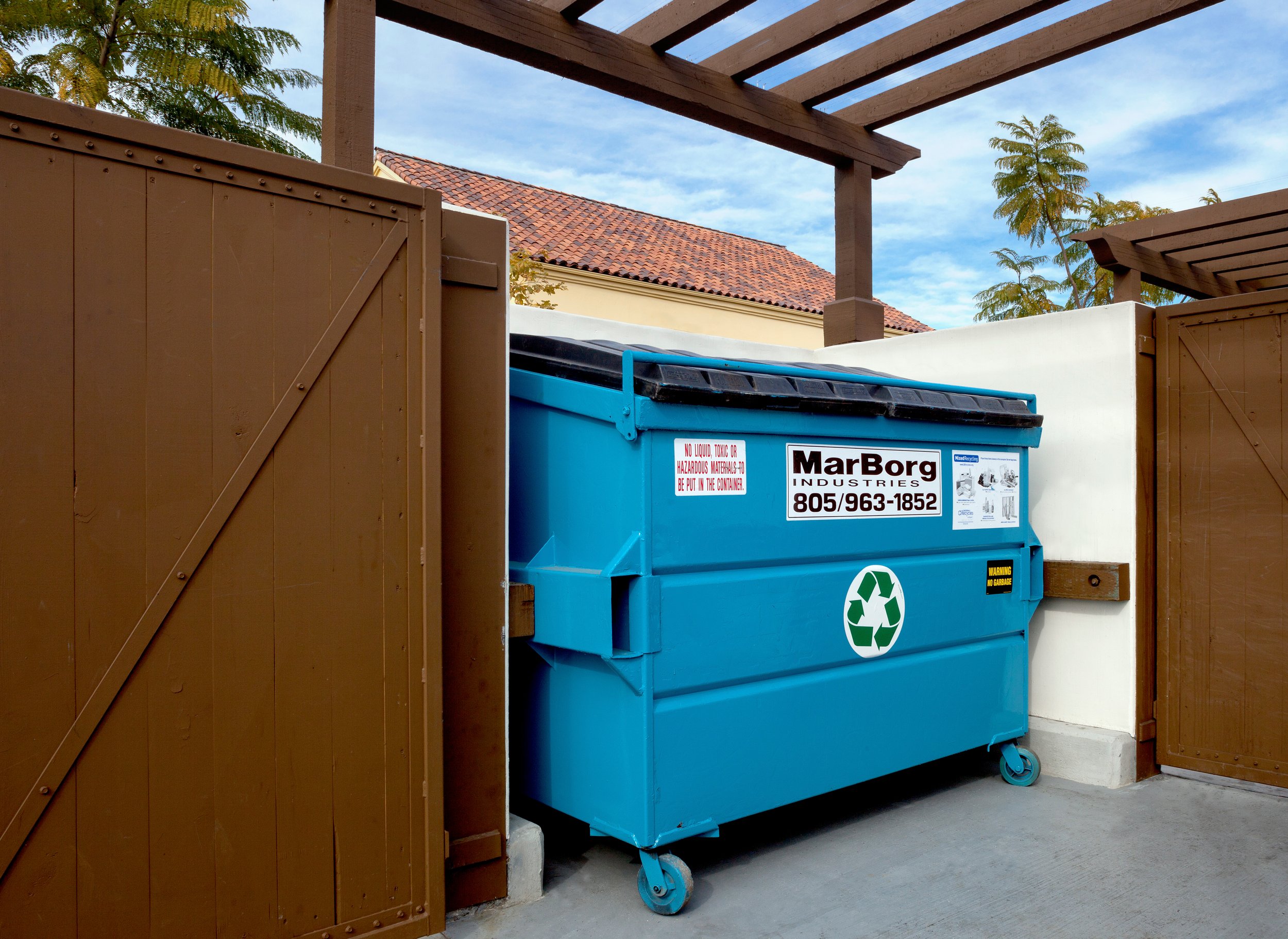 A blue recycling dumpster positioned between brown wooden fences, with a white wall behind it, under a wooden pergola with a clear sky and a house with a red tiled roof in the background.