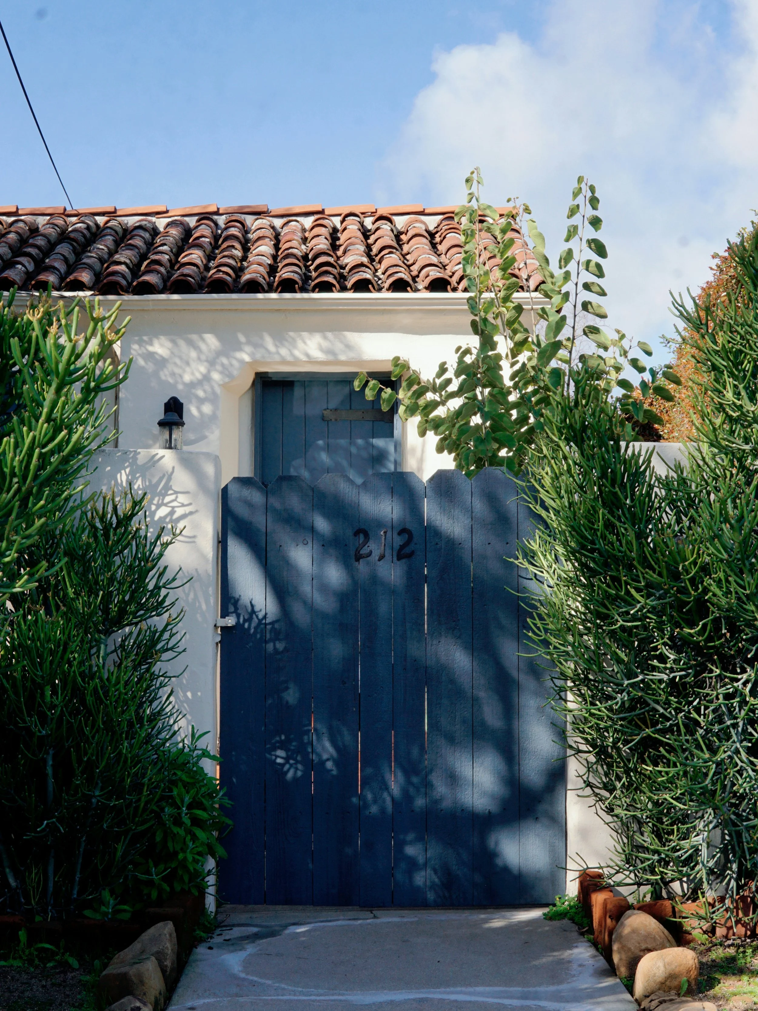 A black wooden gate with the number 212 painted on it, set in a white wall, with lush green plants on each side, a small brick wall at the base, and a tiled roof above against a blue sky.