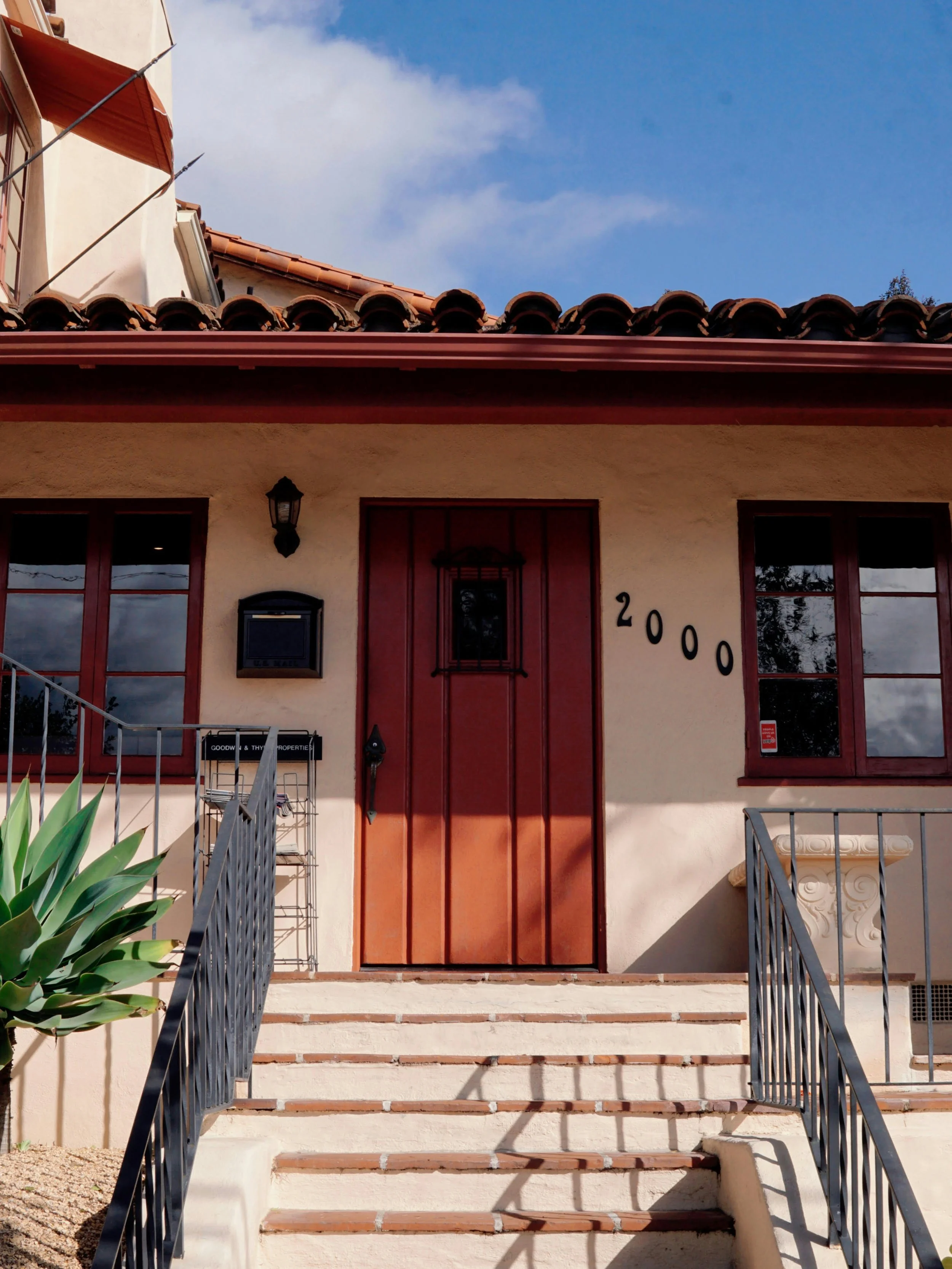 Exterior view of a house with a staircase leading up to a front door, featuring a red door, two windows, a wall-mounted lantern, and a house number 2000, under a blue sky with some clouds. There are plants on the left side and metal railings on both sides of the stairs.