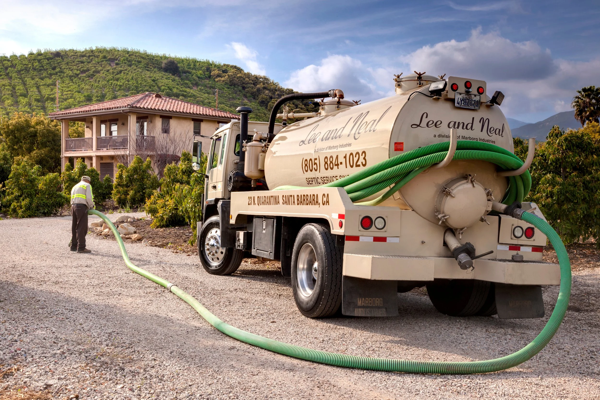 A septic service truck with green hoses getting ready for work next to a house in a residential area with green hills and partly cloudy sky in the background.