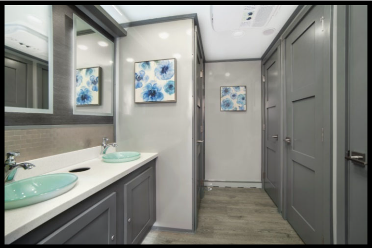 Luxury portable restroom with two mint green sinks, gray cabinetry, a large mirror, floral artwork, and gray doors.