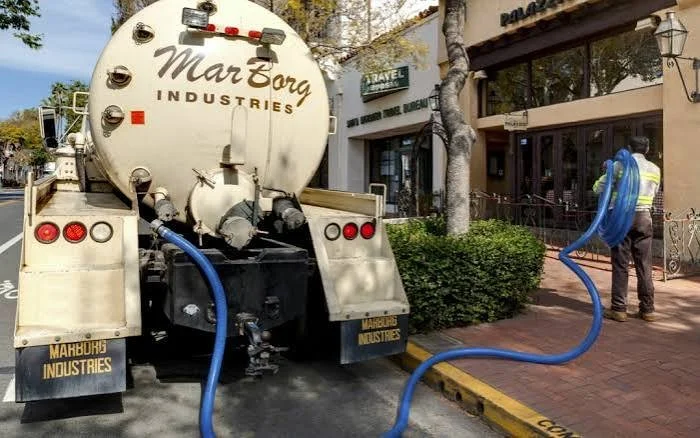 A vacuum truck with blue hoses parked on a city street, with a worker nearby in front of a building.
