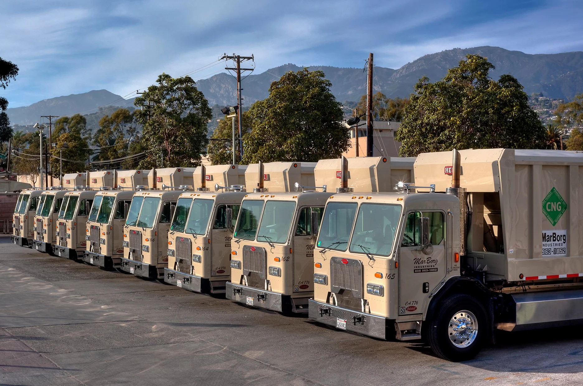 Line of automated side loader trucks with CNG labels parked in outdoor MarBorg lot, with Santa Barbara mountains and trees in the background.