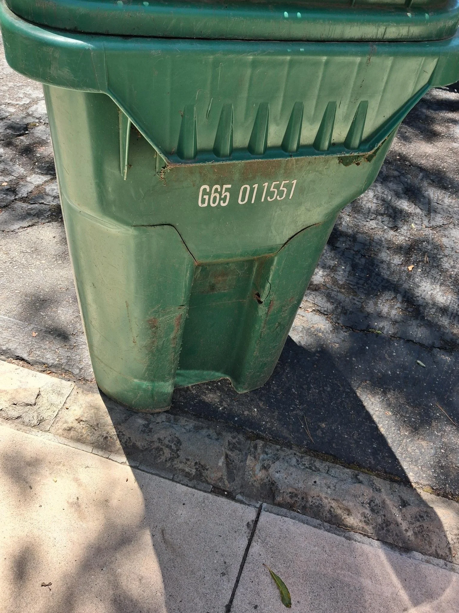 A green outdoor trash bin with a damaged front panel and a serial number 'G65 01155' printed on it, situated on a concrete and asphalt surface with some fallen leaves and shadows.