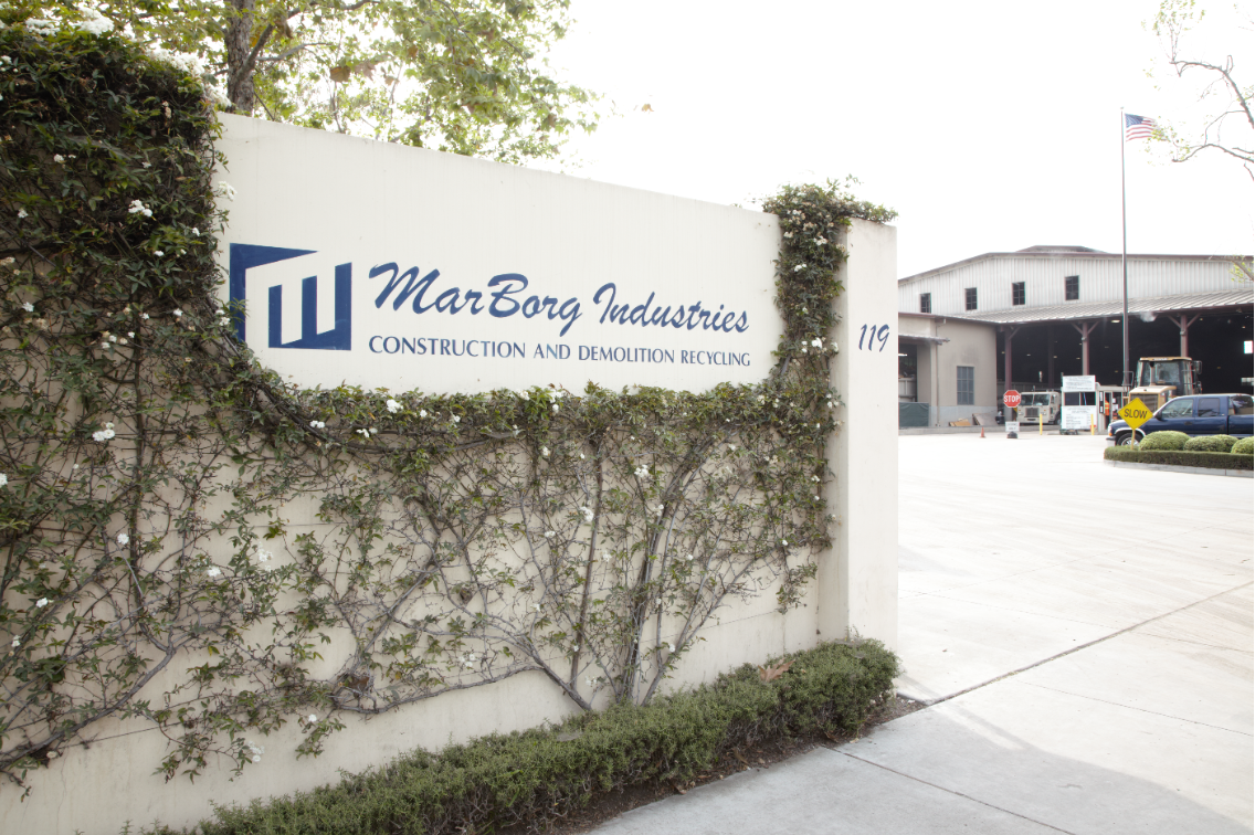 Front entrance to MarBorg Industries Construction and Demolition Recycling facility located on Quarantina Street in Santa Barbara, California. There are trucks in the background and a tall American flag waiving overhead.