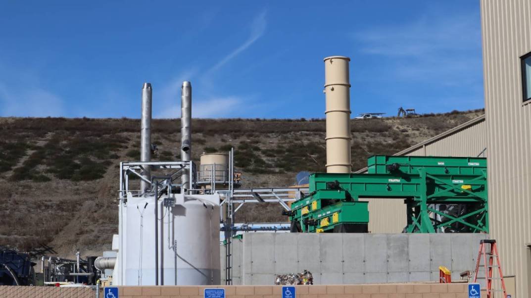 Industrial facility with large green machines, metal pipes, and exhaust stacks against a hillside and blue sky.